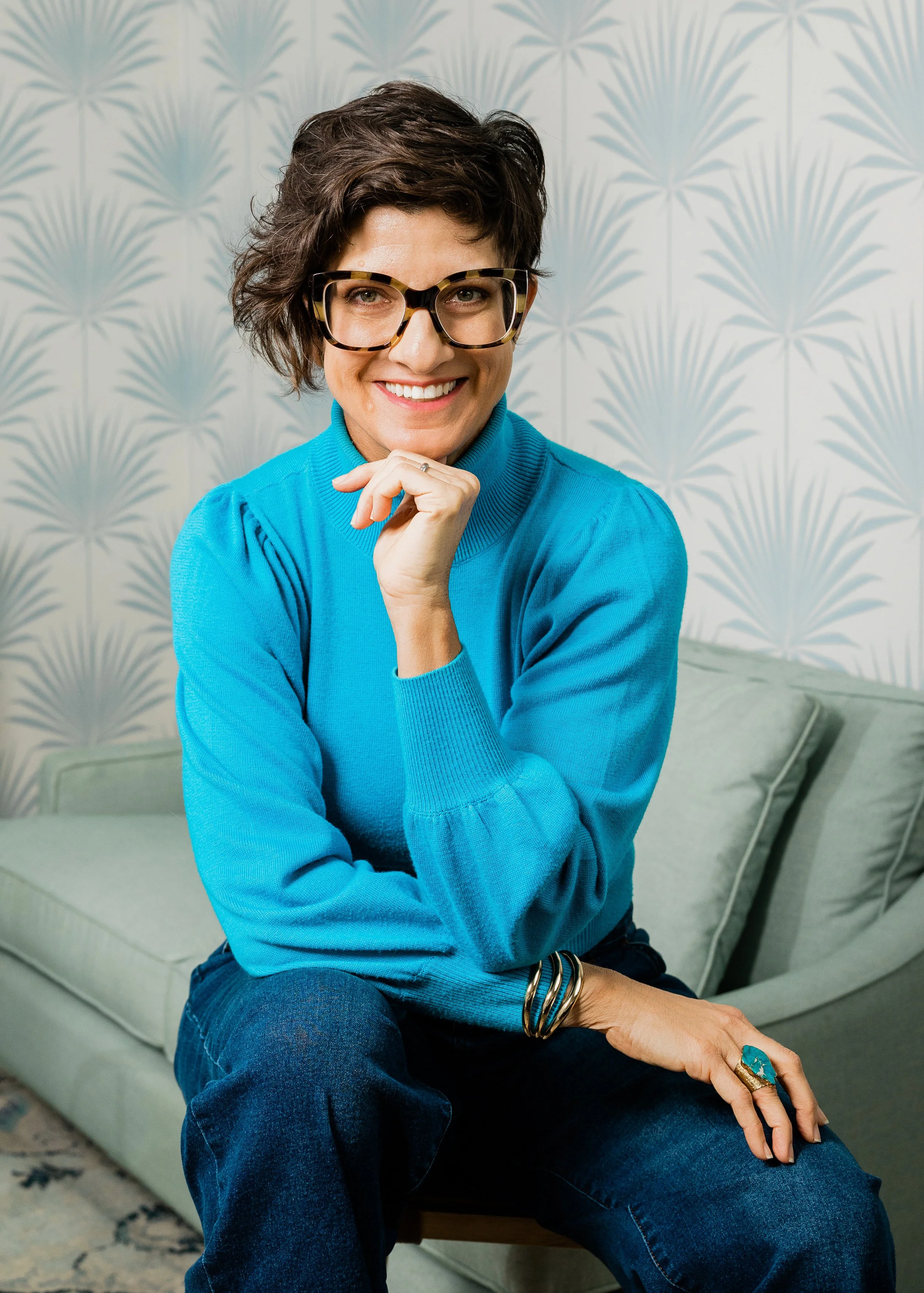 Amy Pezzicara is pictured  with short dark hair and glasses, smiling and sitting on a light-colored sofa with a patterned wallpaper background. She is wearing a bright blue sweater, dark blue jeans, and gold and turquoise jewelry.