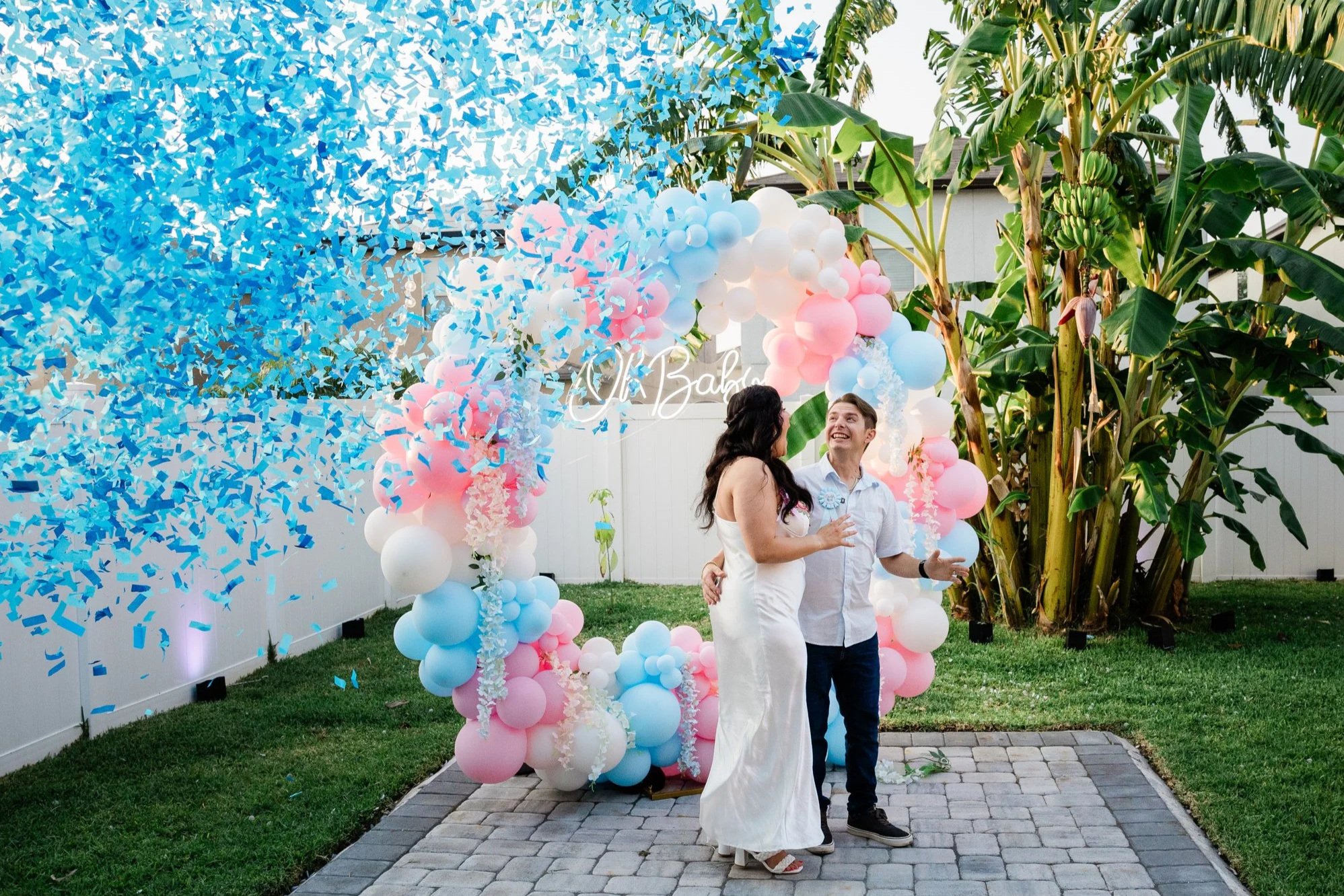 A couple standing in front of balloon decorations at a backyard celebration, with a woman taking a photo, and a large banana plant in the background.
