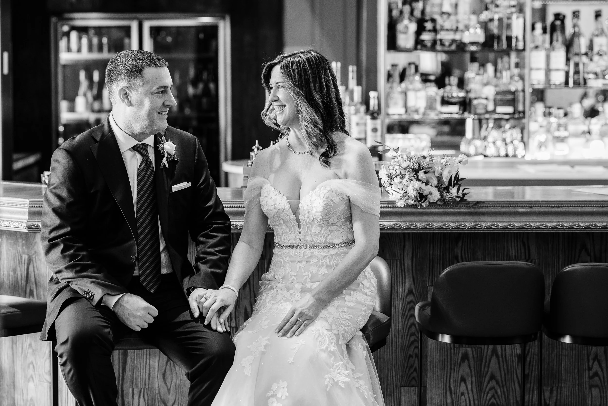 A black and white photo of a bride and groom sitting close together, holding hands and smiling at each other. The groom is wearing a dark suit with a tie, and the bride is in a lace wedding dress with an off-the-shoulder design, seated in front of a 