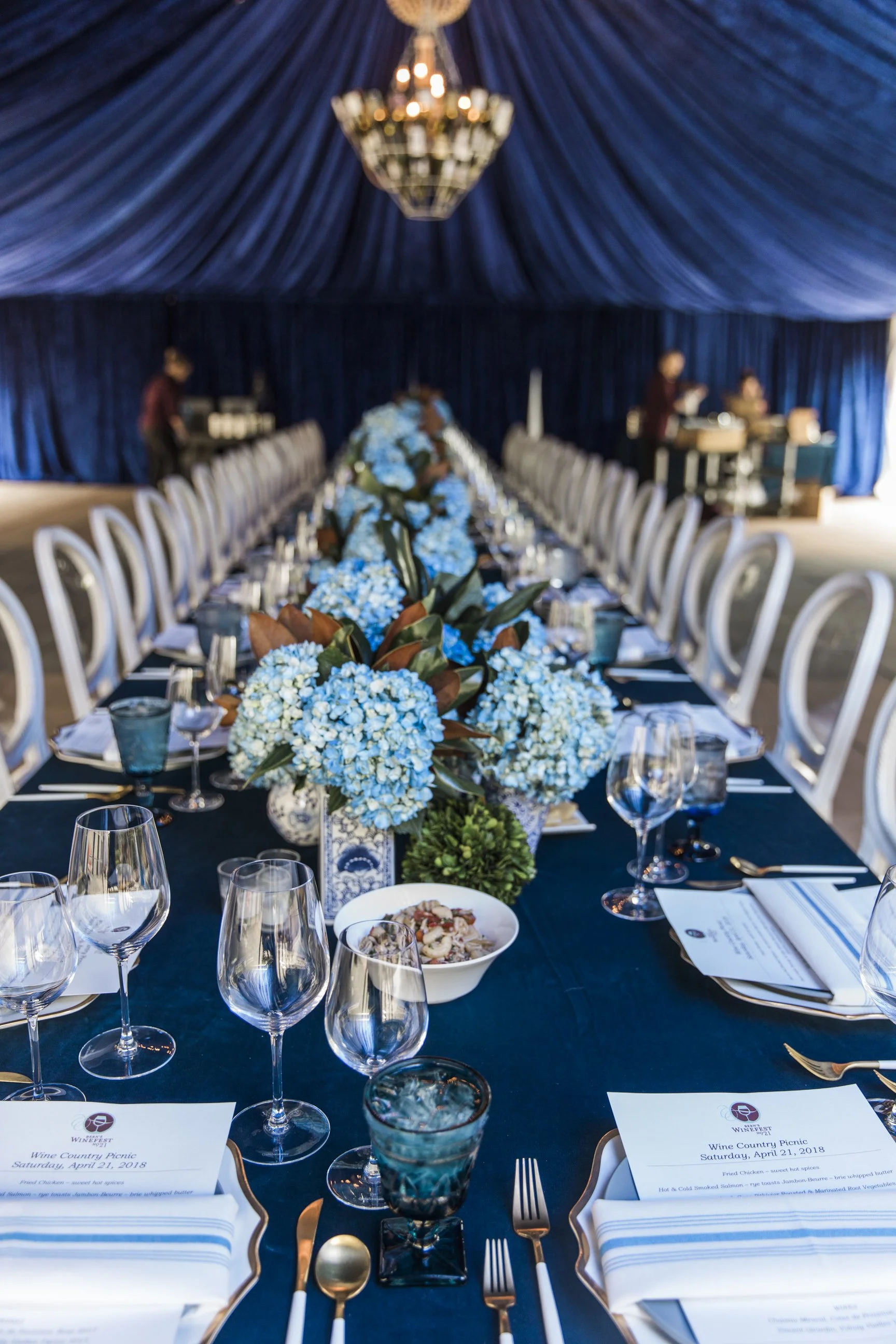Elegant banquet table with white napkins, gold flatware, wine glasses, and a floral centerpiece with blue and white flowers. The setting is under a blue draped ceiling in a large tent, with menu cards and food dishes visible. There are people prepari