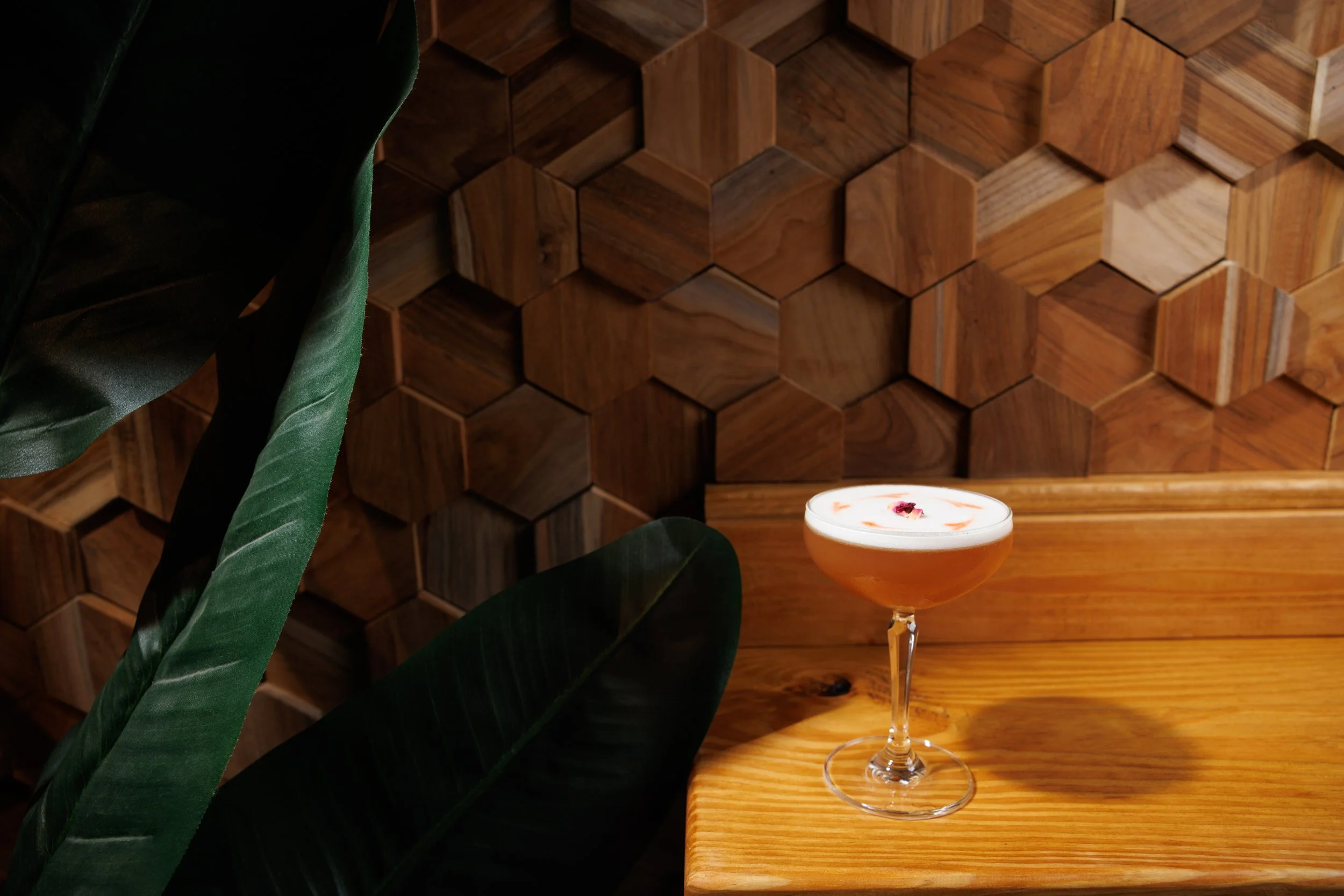 A cocktail glass with a pink drink and garnished with a cherry on top, placed on a wooden table against a wood-paneled wall.