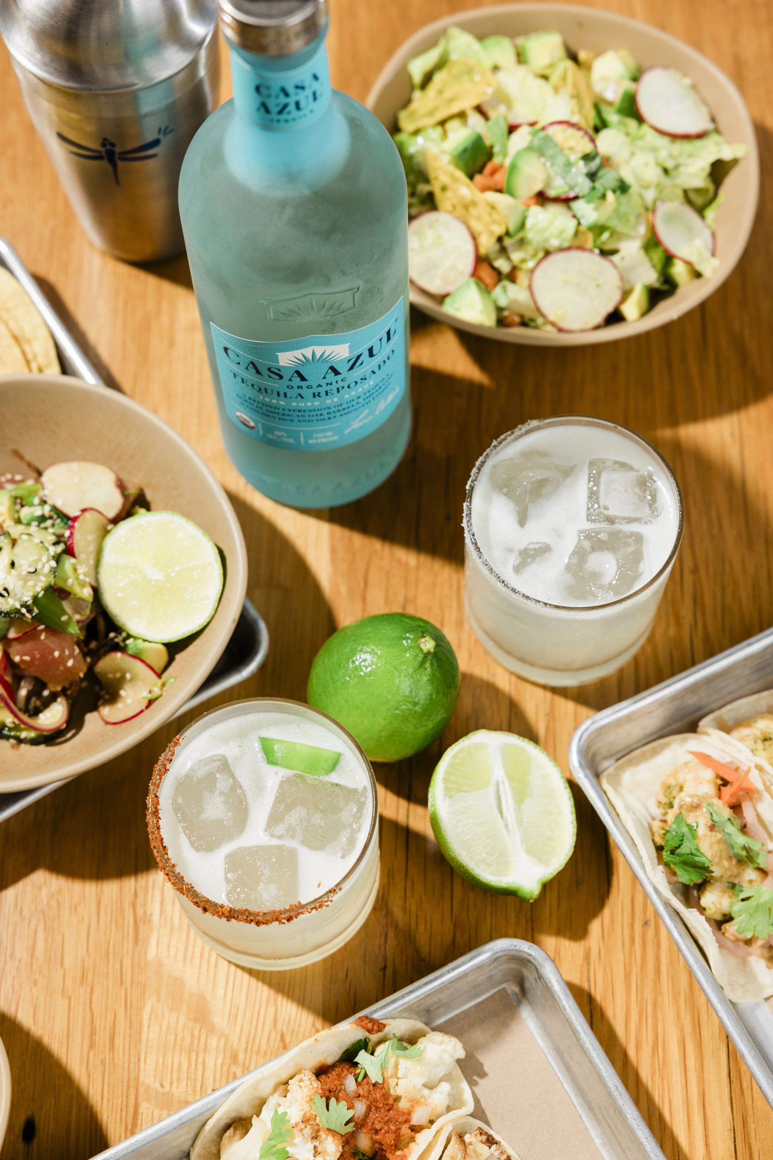 A table with two bottles of Casa Azul organic liquor, two glasses of ice-cold tequila with salted rims, a green lime, two halves of a lemon, a bowl of salad with radishes and avocado, and various small plates of Mexican dishes.