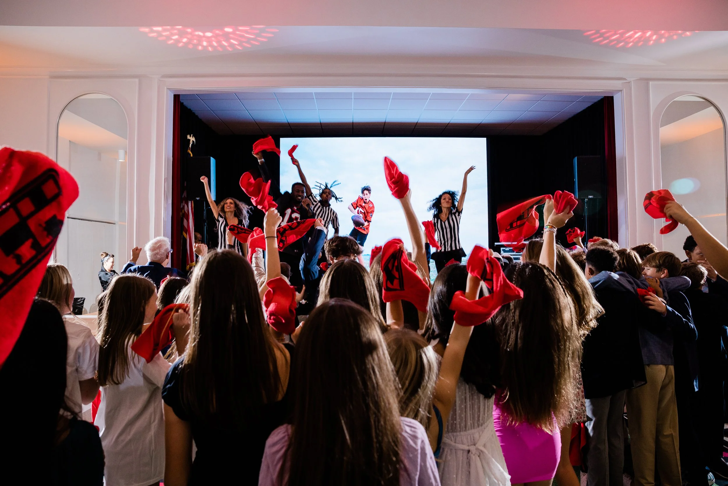 A group of children and adults attending an indoor event, many holding red bandanas, with some people on stage dressed in football jerseys and referees, on a stage with a large screen behind them.