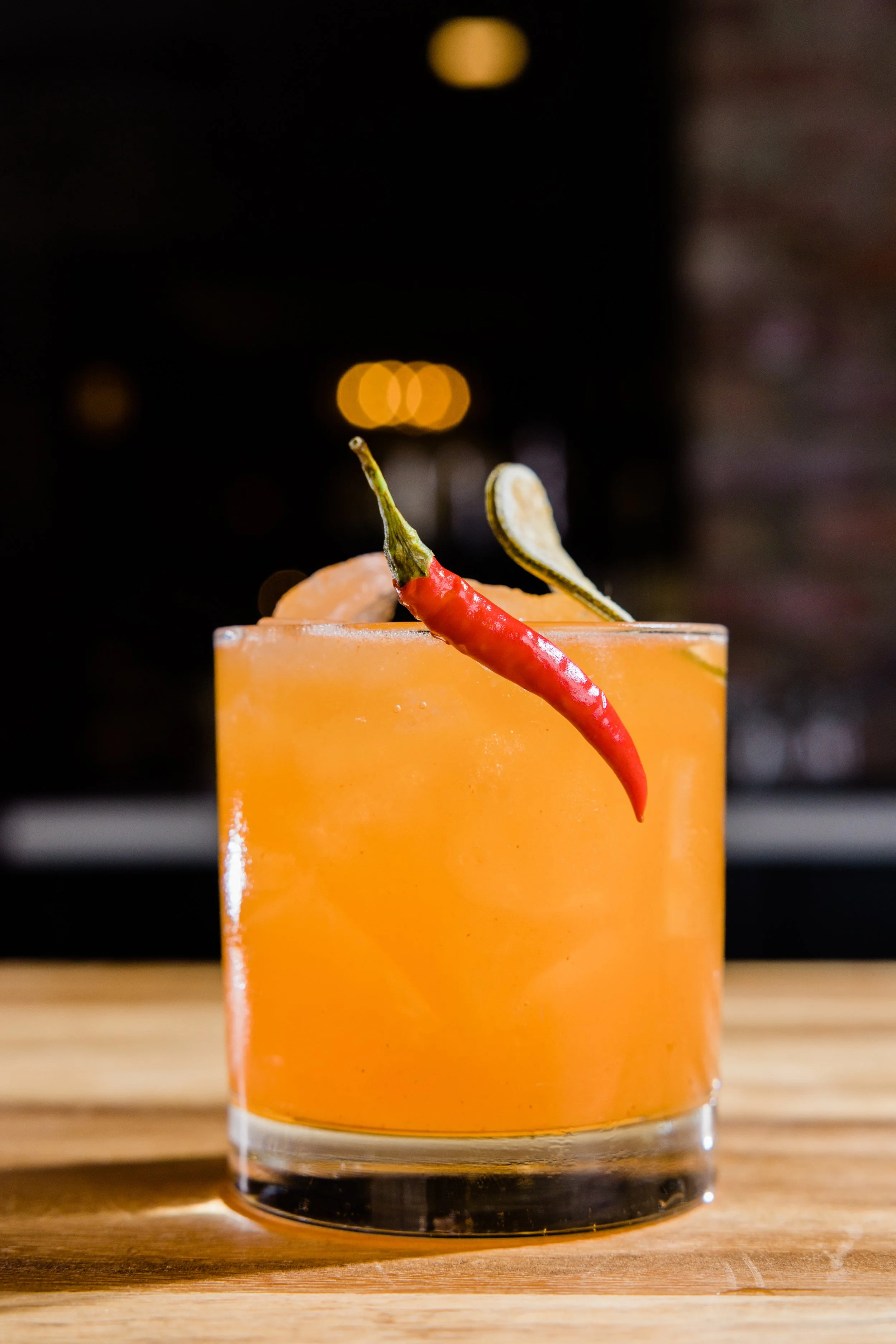 Close-up of a bright orange cocktail in a clear glass, garnished with a red chili pepper and a slice of lemon on a wooden surface with a blurred dark background.
