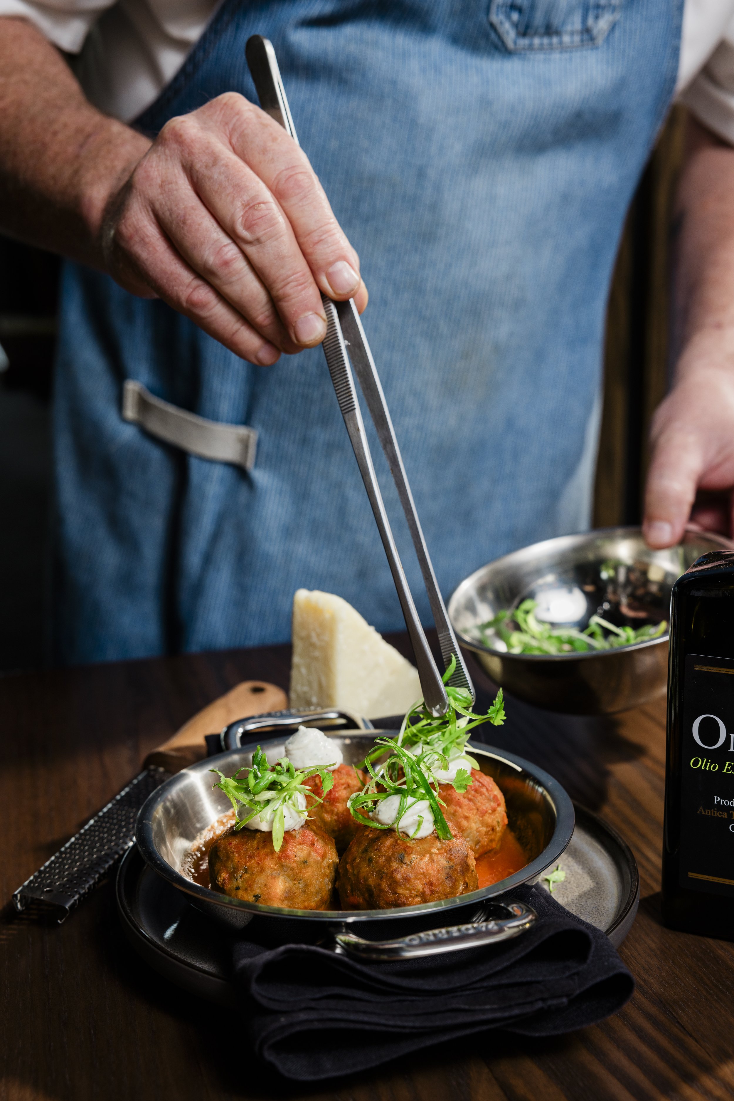 Person in a blue apron preparing a dish of meatballs garnished with herbs, with a cheese wedge and salad in the background.