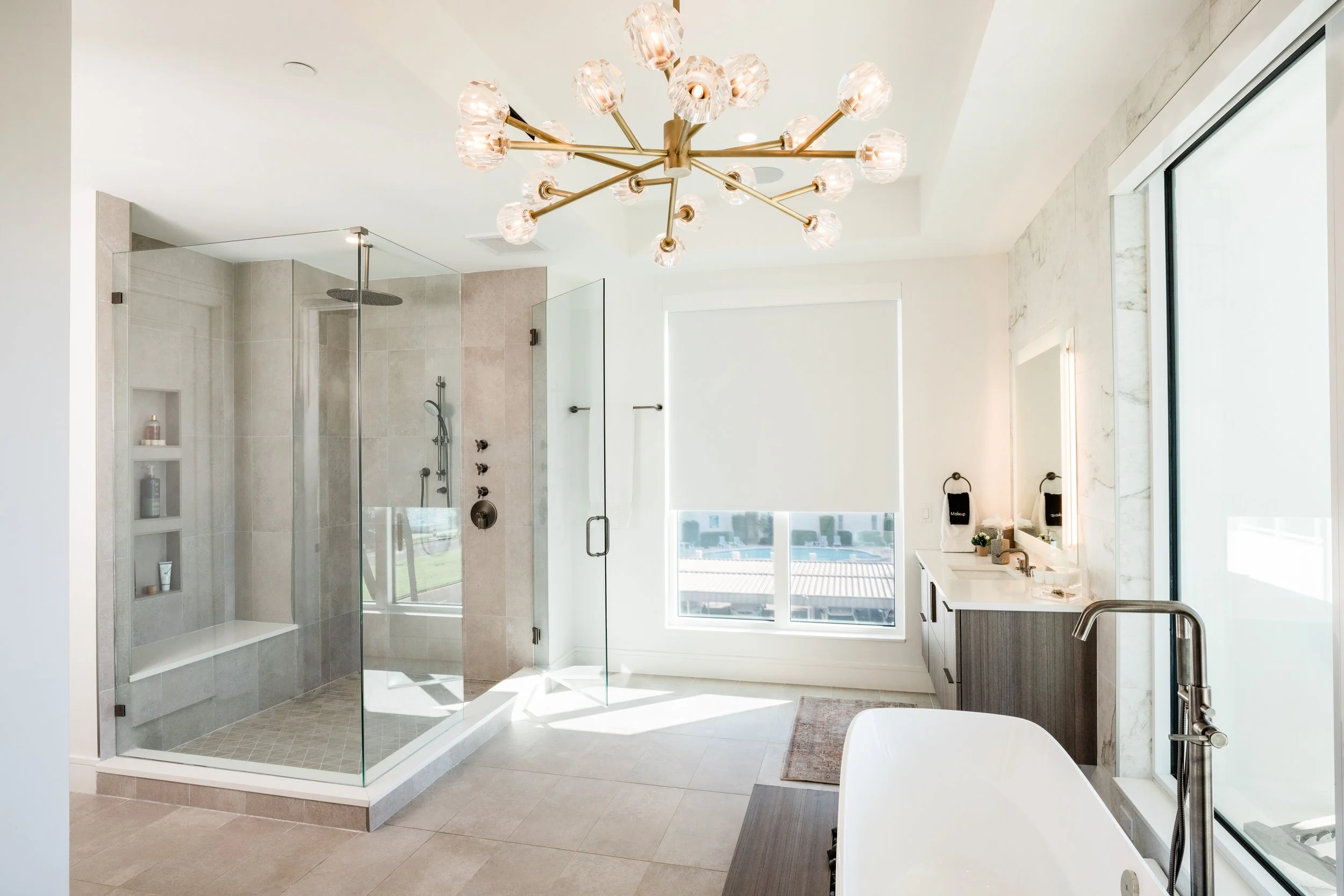 Modern bathroom with a glass-enclosed walk-in shower, large window, and a freestanding bathtub, featuring a gold chandelier with multiple bulbs hanging from the ceiling. Interior design by jamie finley & co.