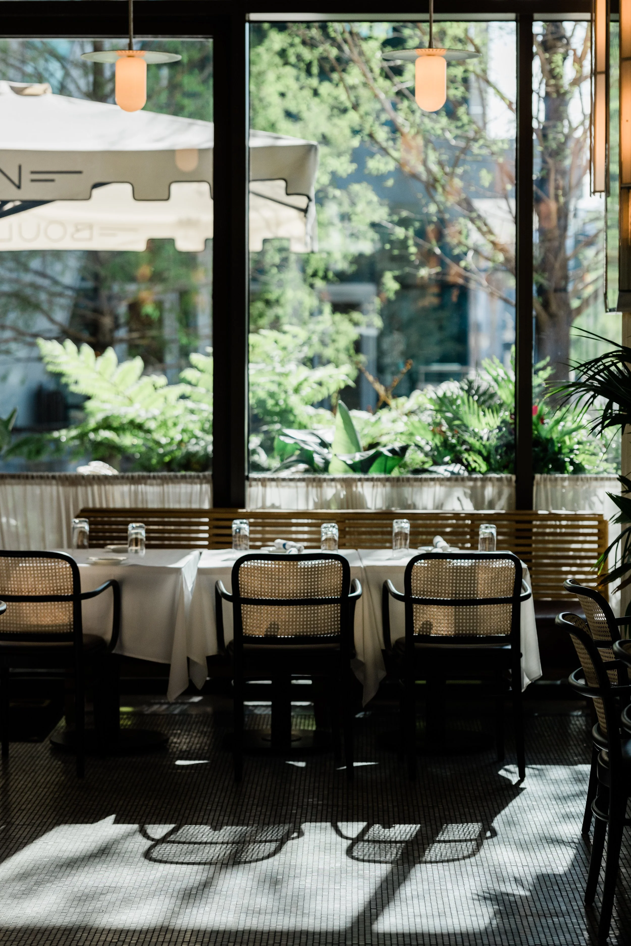 Empty restaurant table with white tablecloth, glasses, and napkins, facing large windows with outdoor greenery visible outside.