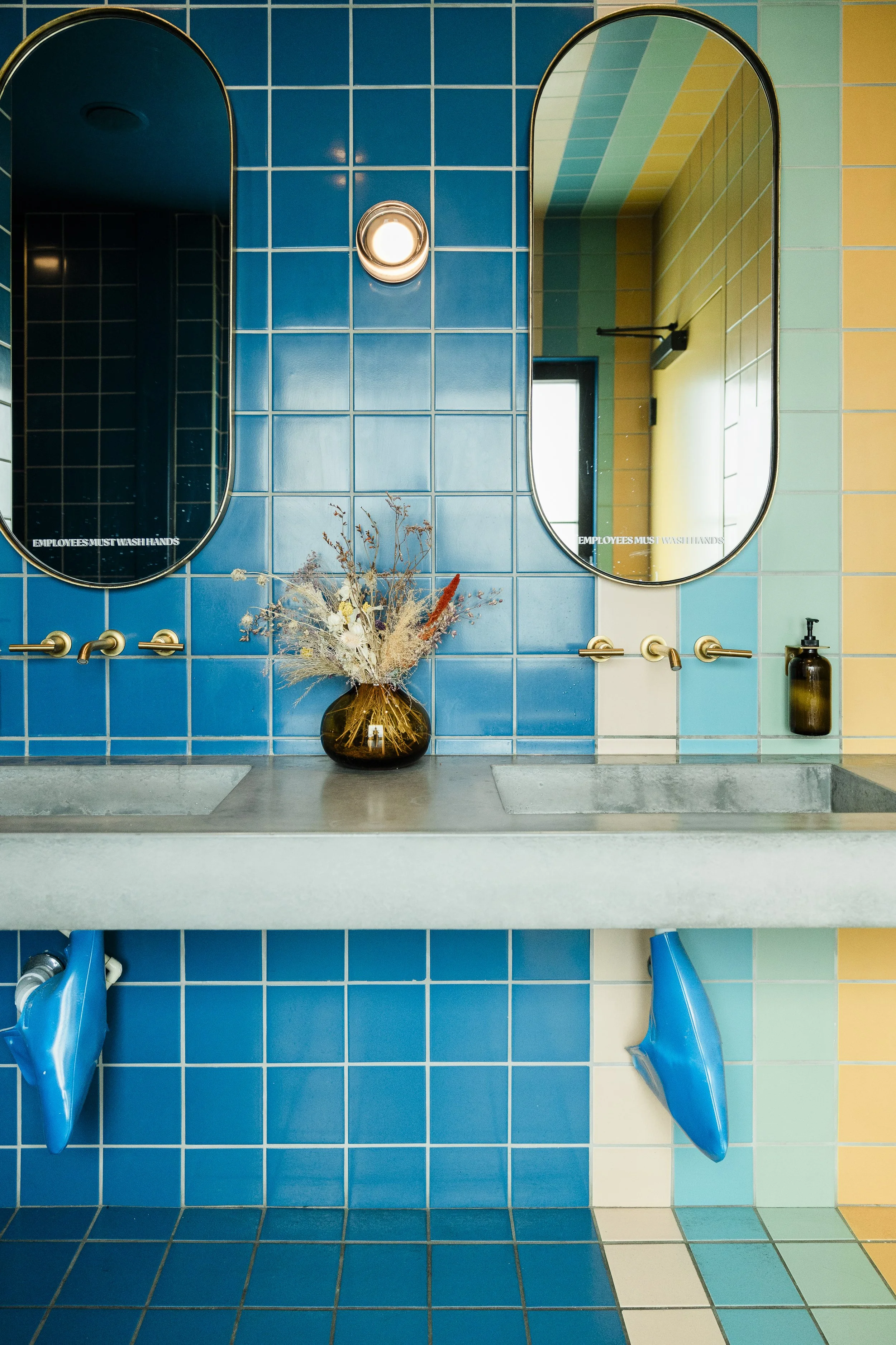 A colorful bathroom with a blue tiled wall, two rectangular mirrors, a vase with dried flowers, a soap dispenser, and blue hand dryers. Willa's restroom 70's retro.