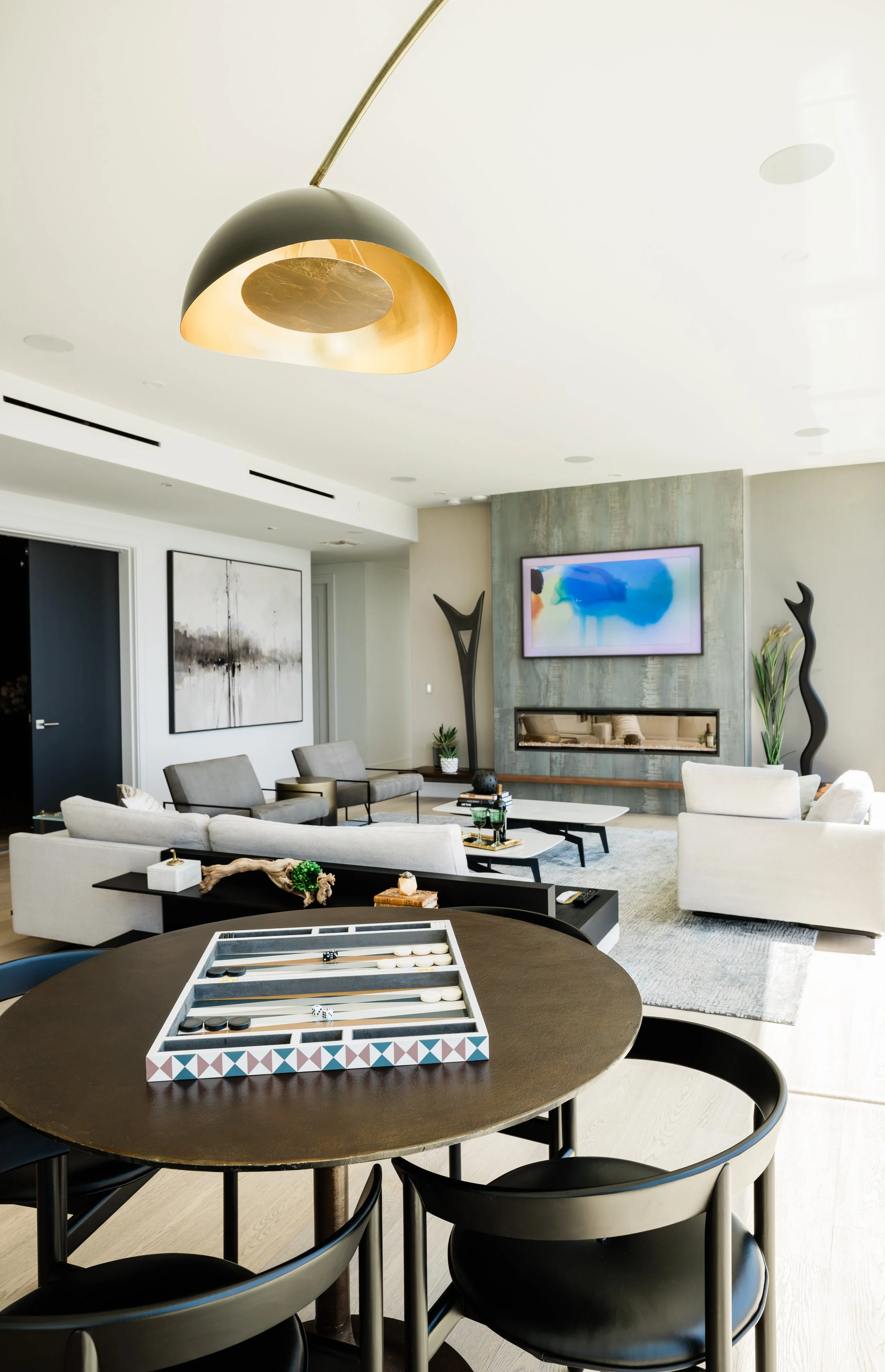 Modern living room with white sofas, a flat-screen TV, abstract artwork, and decorative sculptures. A black dining table with chairs has a backgammon game set on it, and a circular table with a board game is visible in the foreground. The room featur