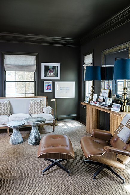 Living room with dark green walls, two large windows with white blinds, a white sofa with patterned pillows, two gray marble side tables, a brown leather lounge chair with an ottoman, a wooden console table with blue lamps, and framed pictures on the