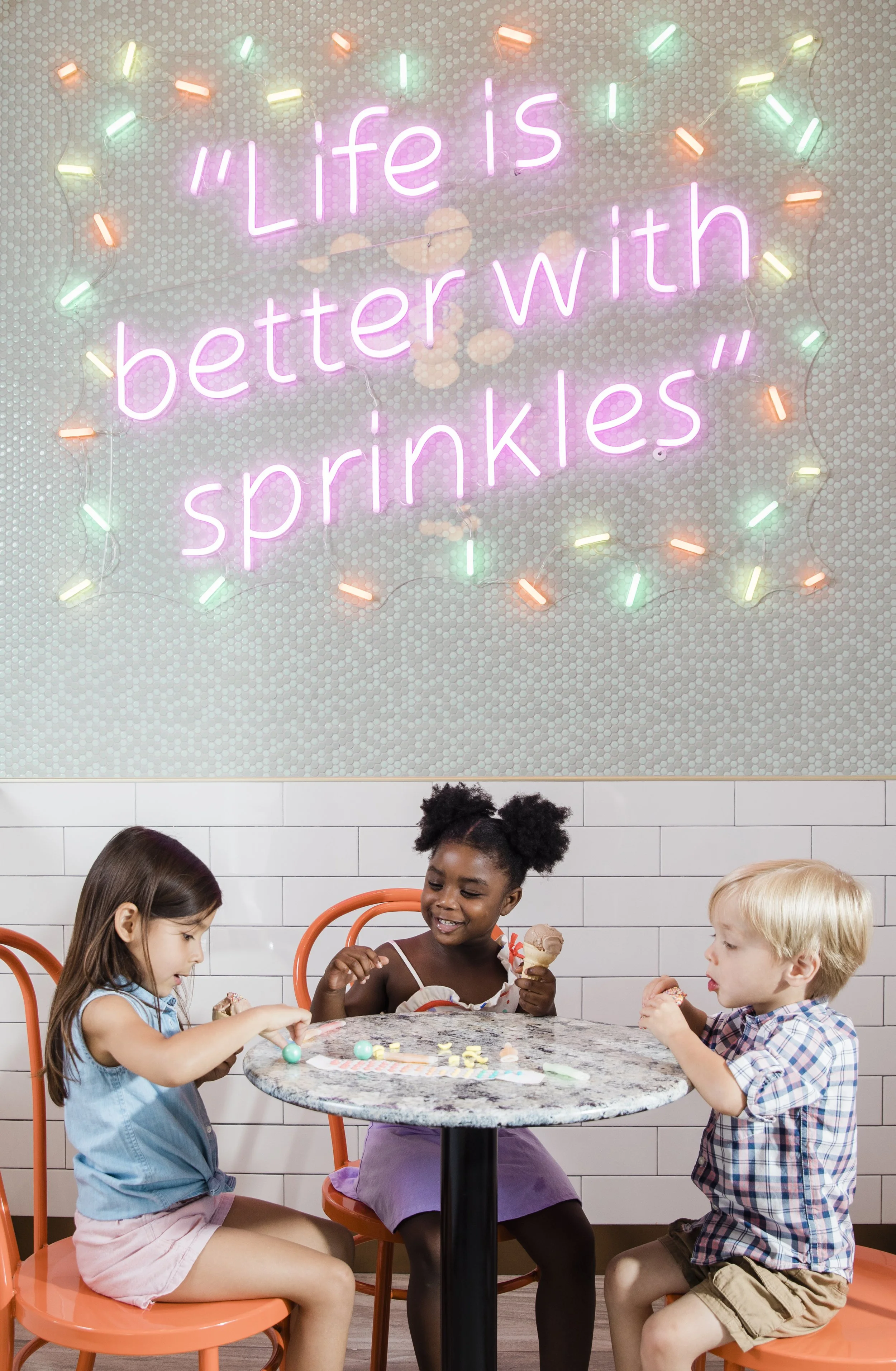 Three kids sit around a small marble table eating ice cream and candy. Neon sign in the background reads, "Life is better with sprinkles". Location was Little Midway ice cream shop at the Epicurean Hotel.
