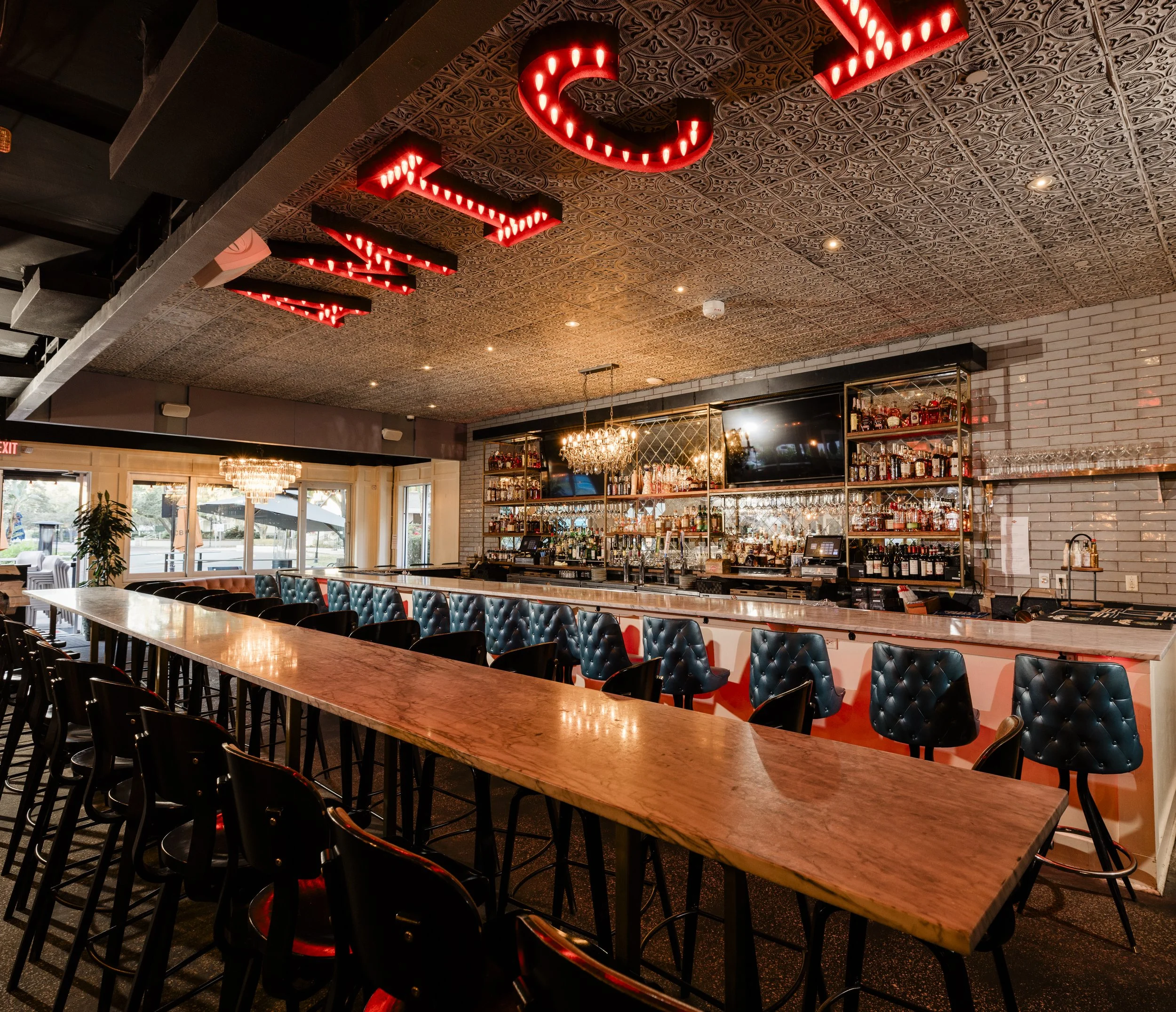 Interior of a bar or restaurant with a long wooden table, black chairs with tufted blue upholstery, a bar area with shelves of liquor bottles, and a ceiling decorated with ornate tin tiles and illuminated red marquee-style letters spelling 'TIKI'.