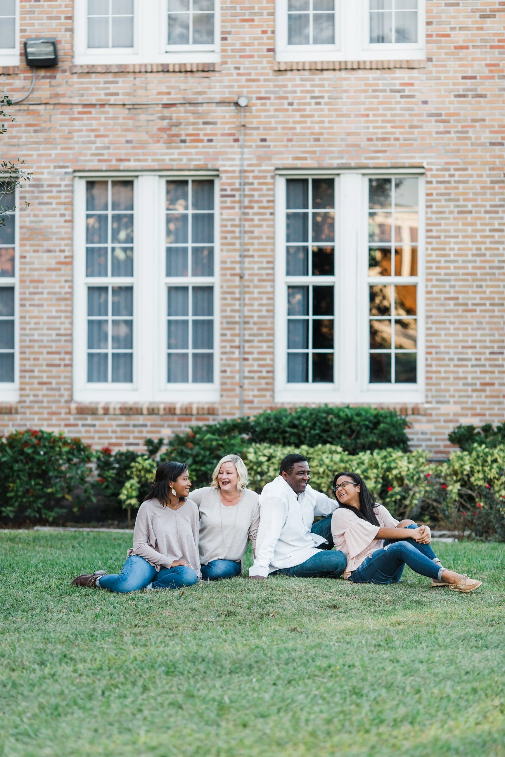 Four people sitting on the grass in front of a brick building, smiling and talking, with bushes and flowers behind them.