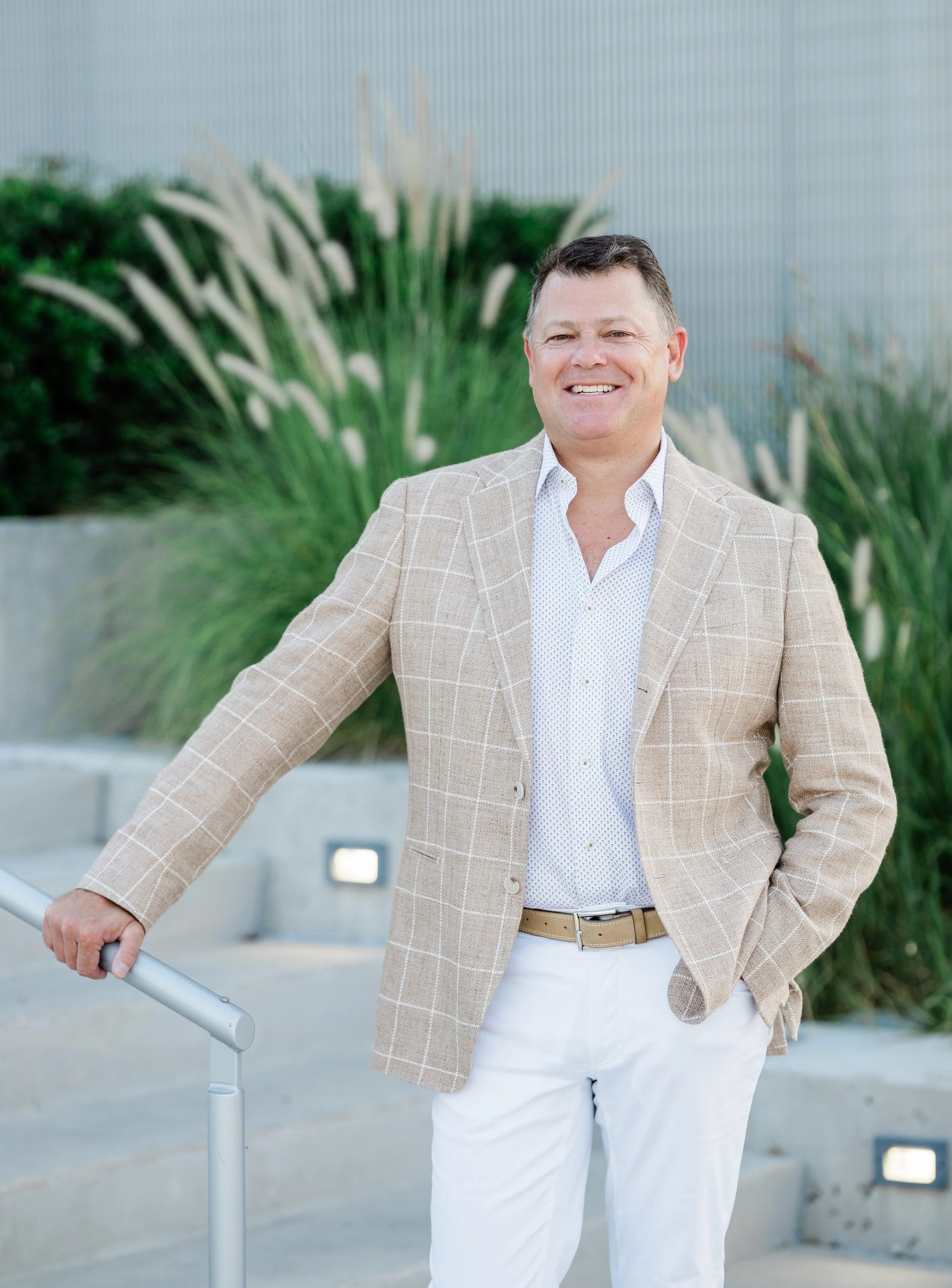 A smiling man in a beige checkered blazer, white shirt, and white pants standing outdoors near a metal railing with greenery in the background.
