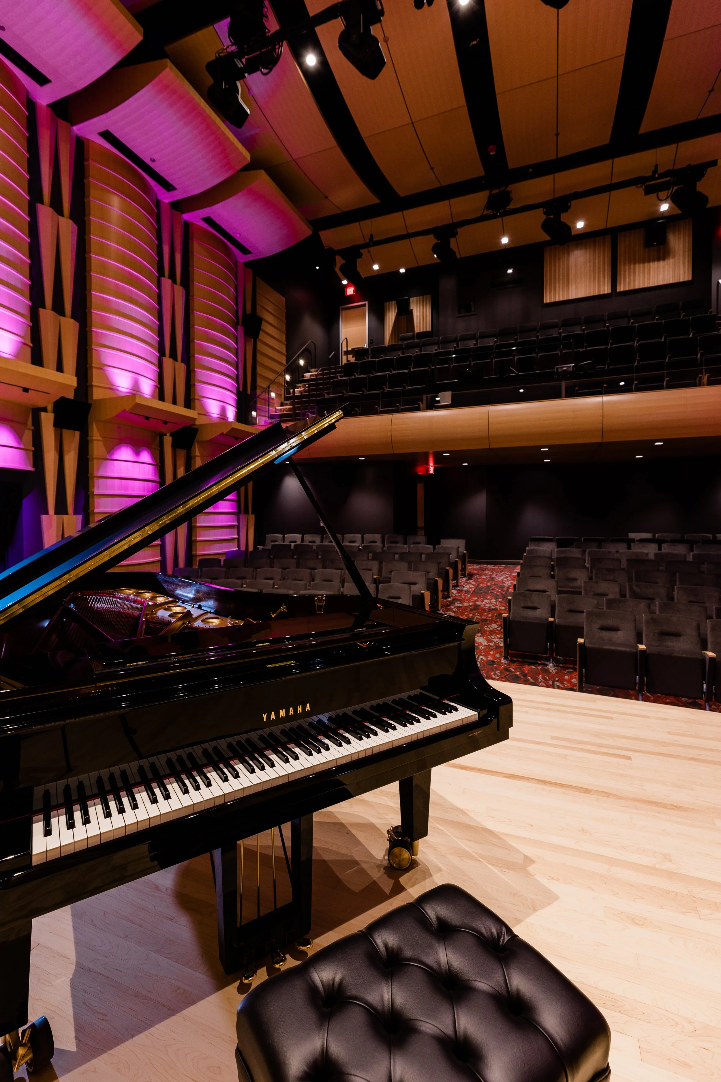 An auditorium with a grand piano on stage, surrounded by rows of seating. The room features wooden paneling, black walls, and purple lighting accents.