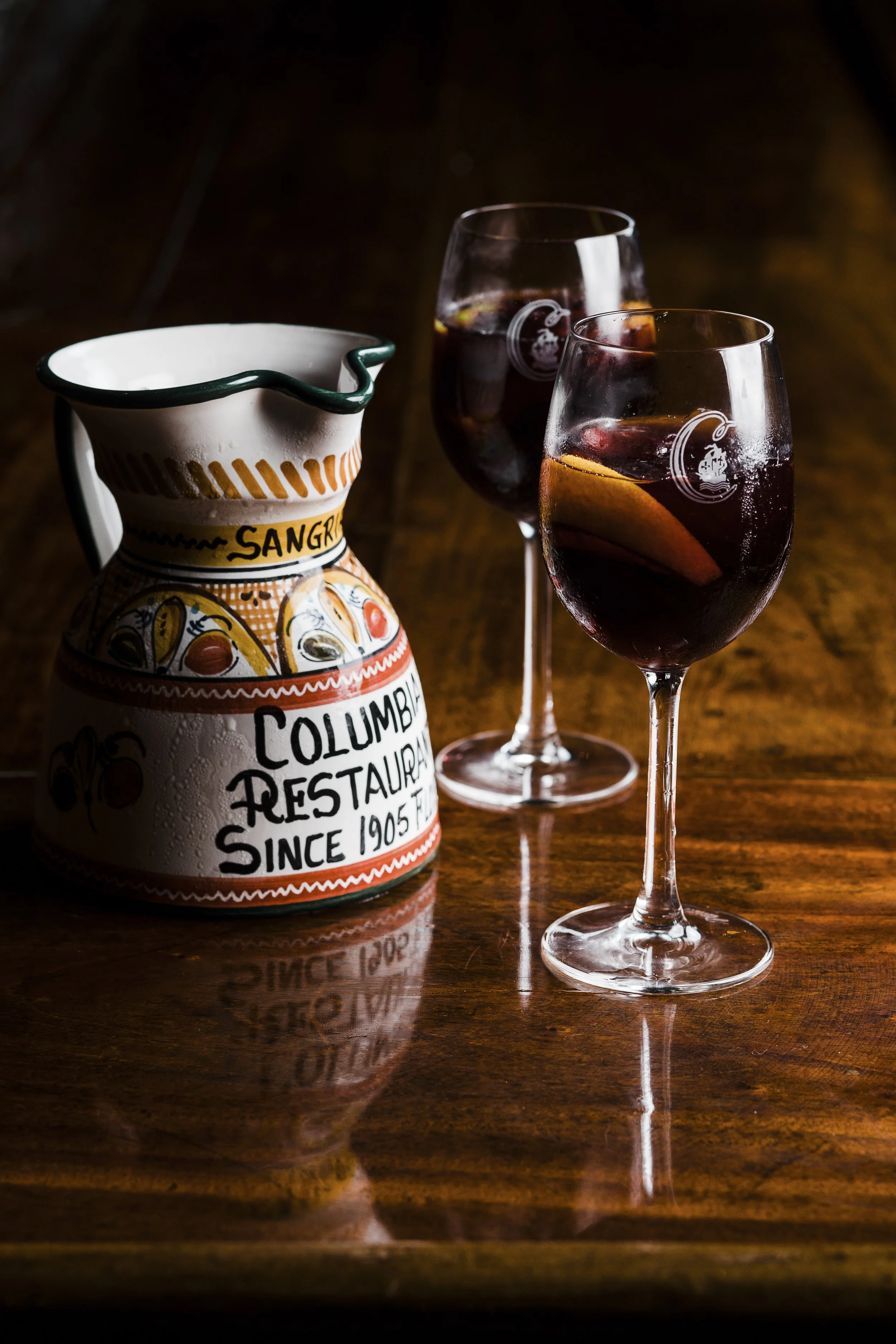 Two glasses of red sangria with orange slices and ice on a wooden table, next to a colorful ceramic pitcher with 'Columbia Restaurant Since 1905' written on it.