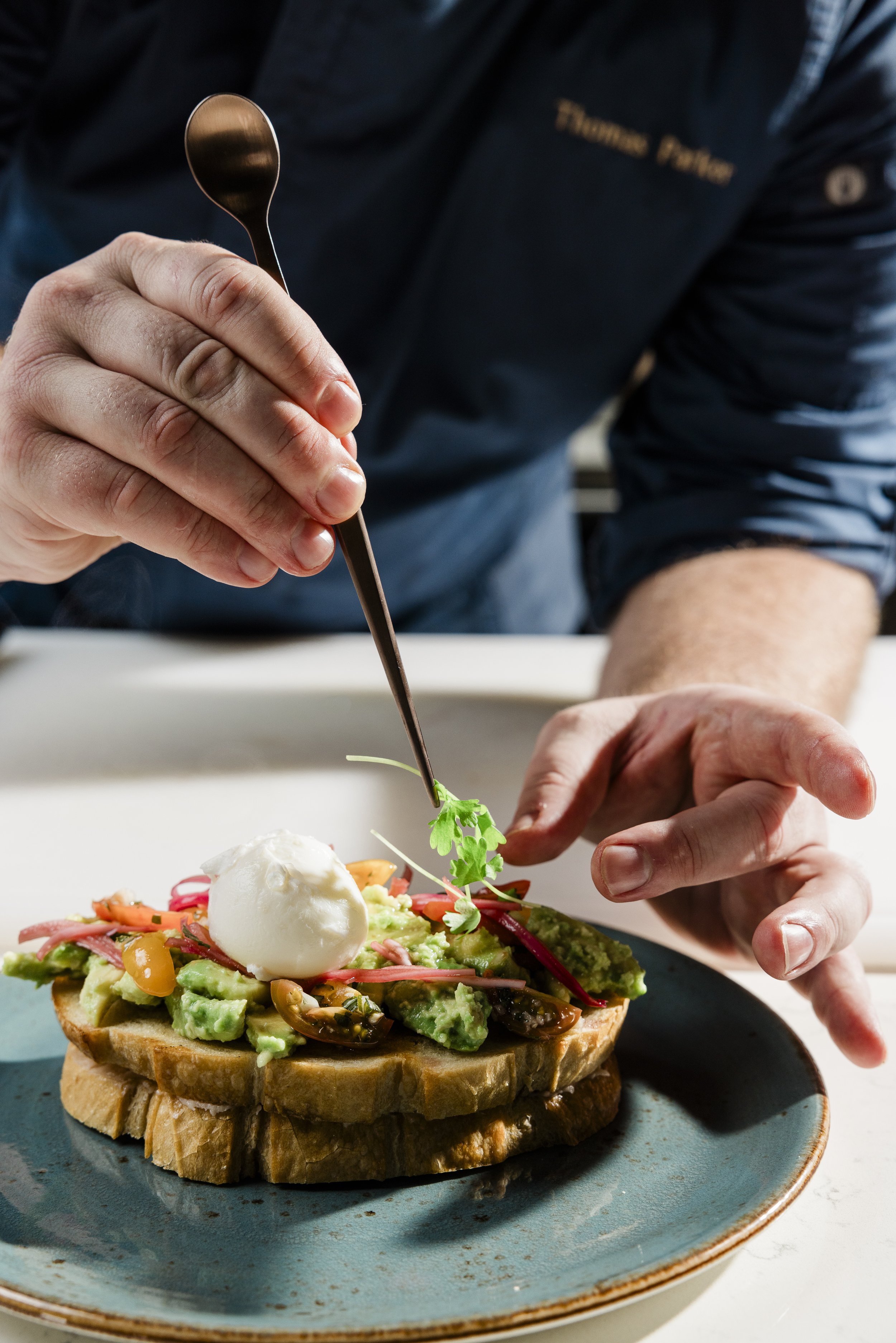 A person assembling a gourmet open-faced sandwich with mashed avocado, cherry tomatoes, pickled onions, and a dollop of sour cream or yogurt on a toasted bread.