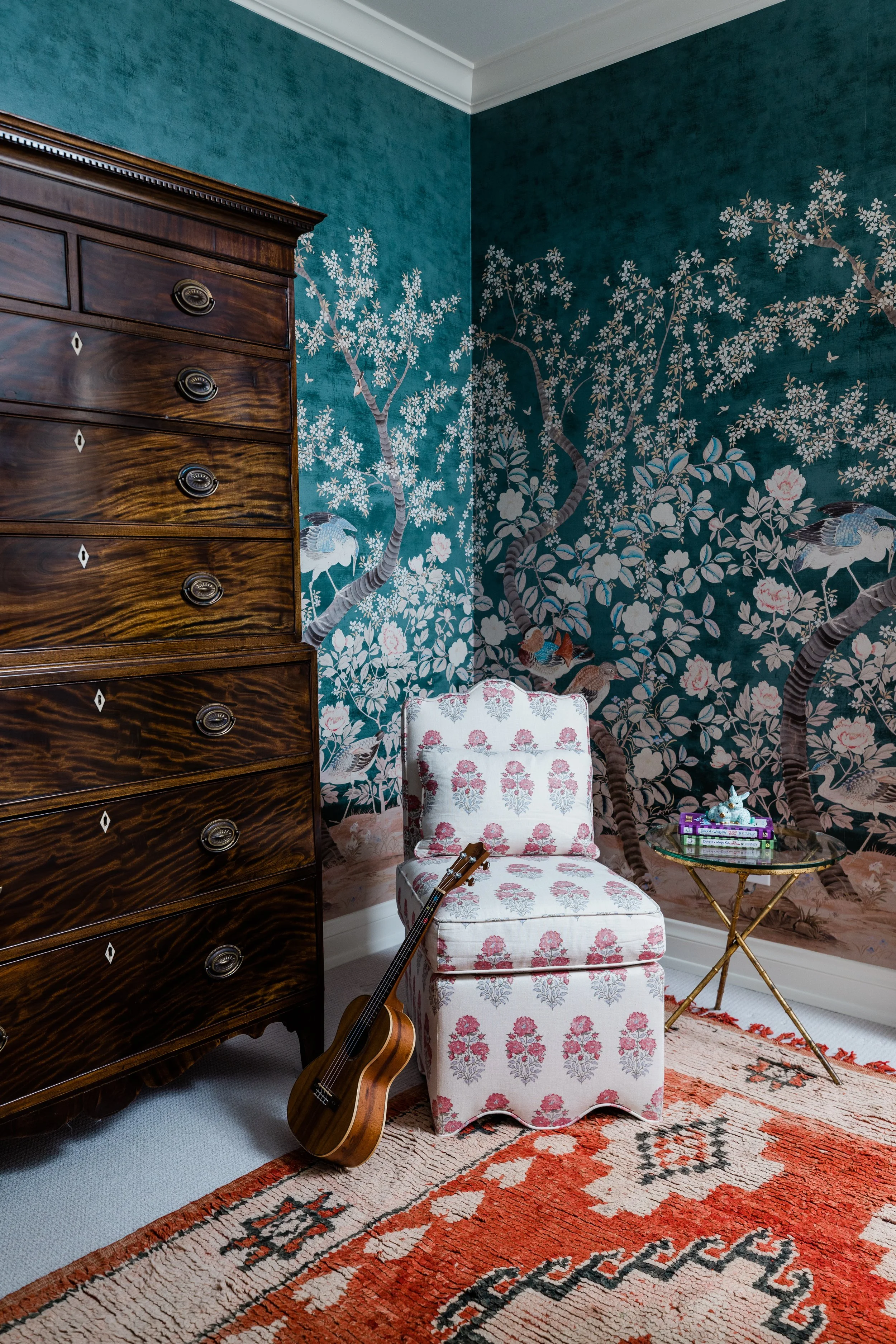 A corner of a room featuring a dark wood dresser, a floral upholstered chair with a guitar leaning against it, a gold metal side table with a stack of books and decorative items, a colorful rug, and a large mural with trees, birds, and flowers painte