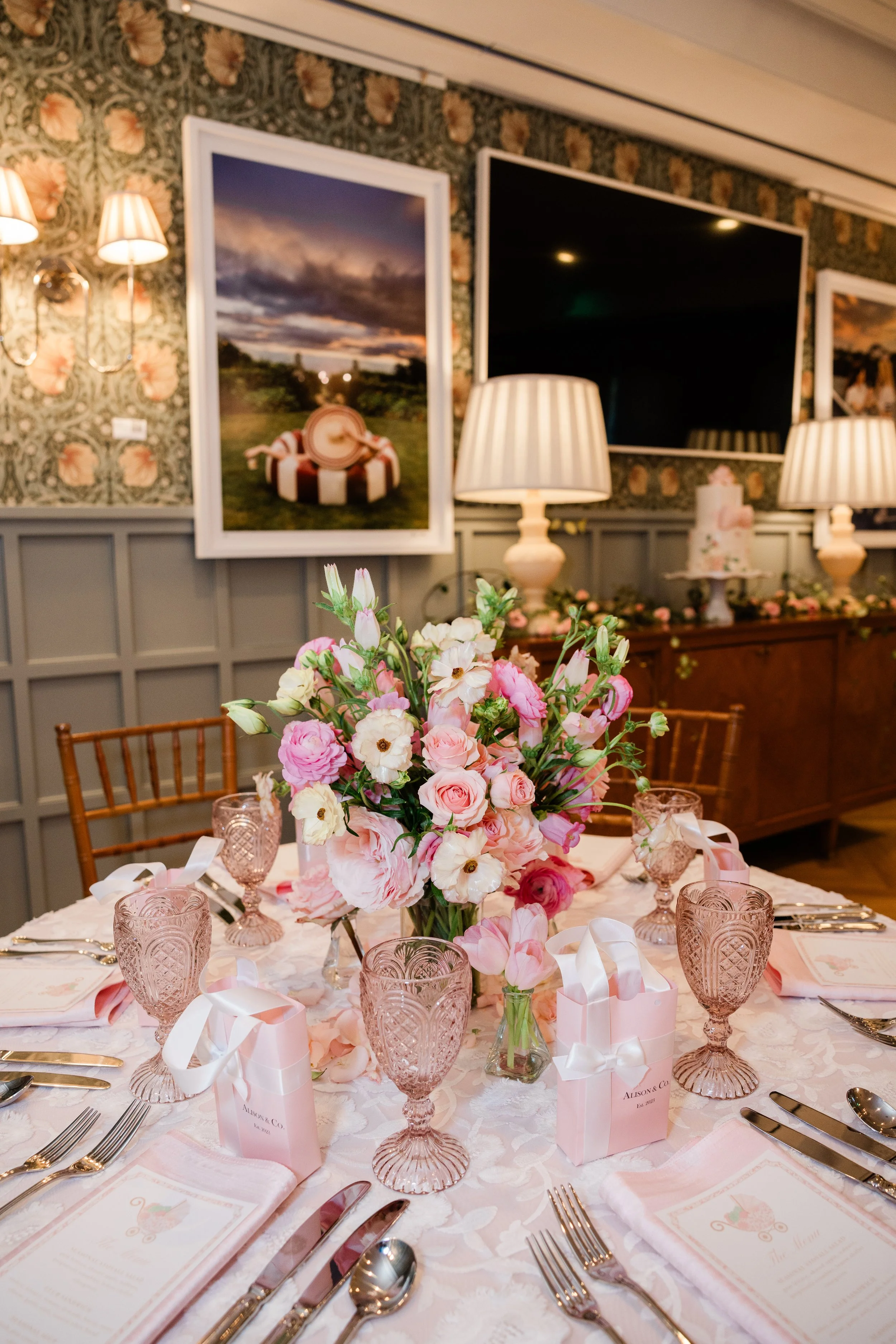 A formal dining table set with pink and white floral centerpiece, pink glasses, menus, silverware, and pink gift bags, surrounded by chairs with a floral wallpaper and framed pictures on the wall. The Stovall House Garden Room.