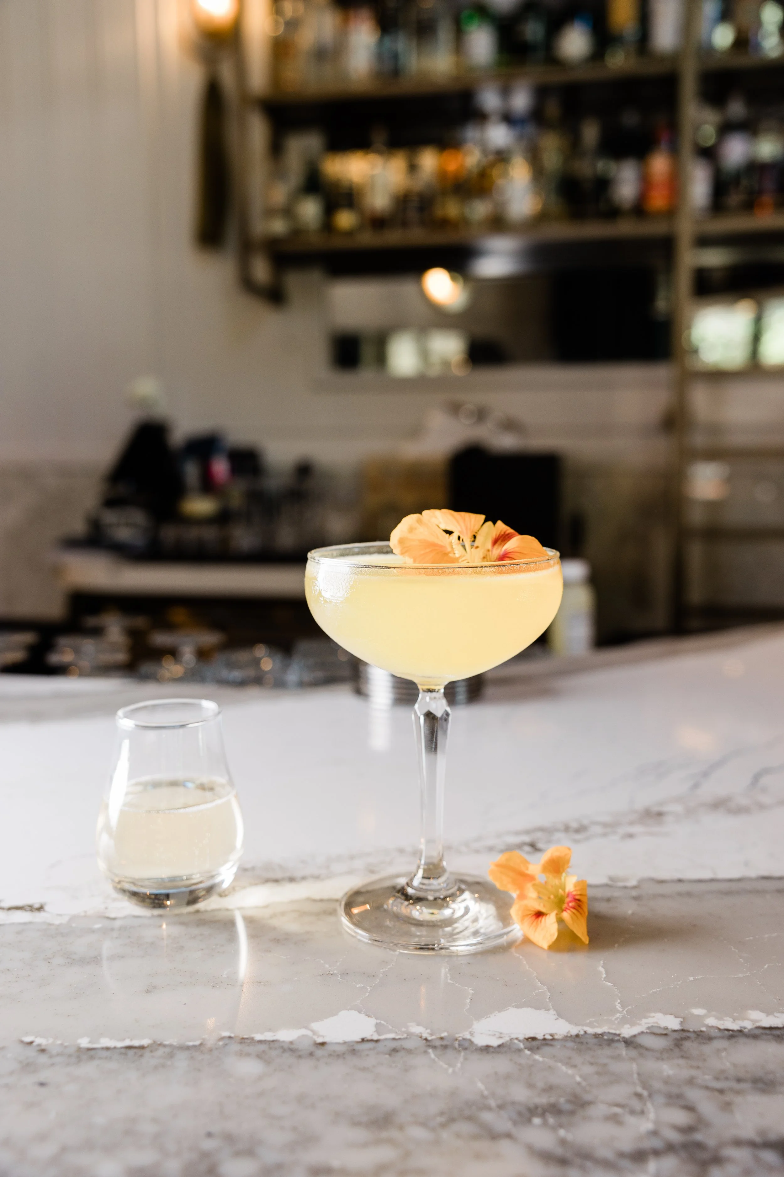 A cocktail glass filled with a yellow drink, garnished with a peach-colored flower, on a marble bar counter with a small glass of clear liquid beside it. Blurred bar shelves with bottles are in the background.