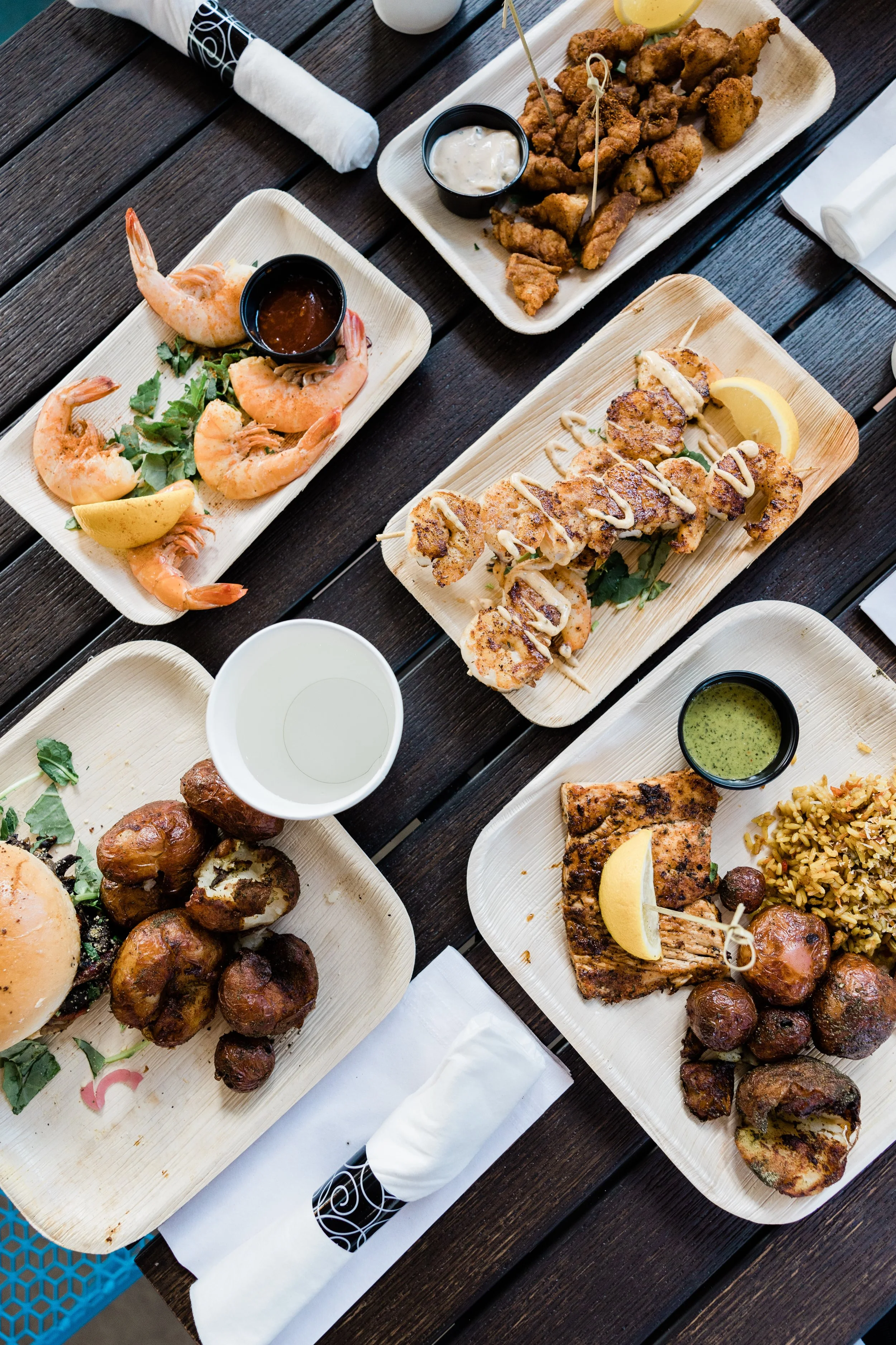 Assorted dishes including shrimp with dipping sauce, fried chicken tenders with lemon, grilled fish with lemon and sauce, roasted potatoes, and rice, all served on bamboo trays at a dining table.