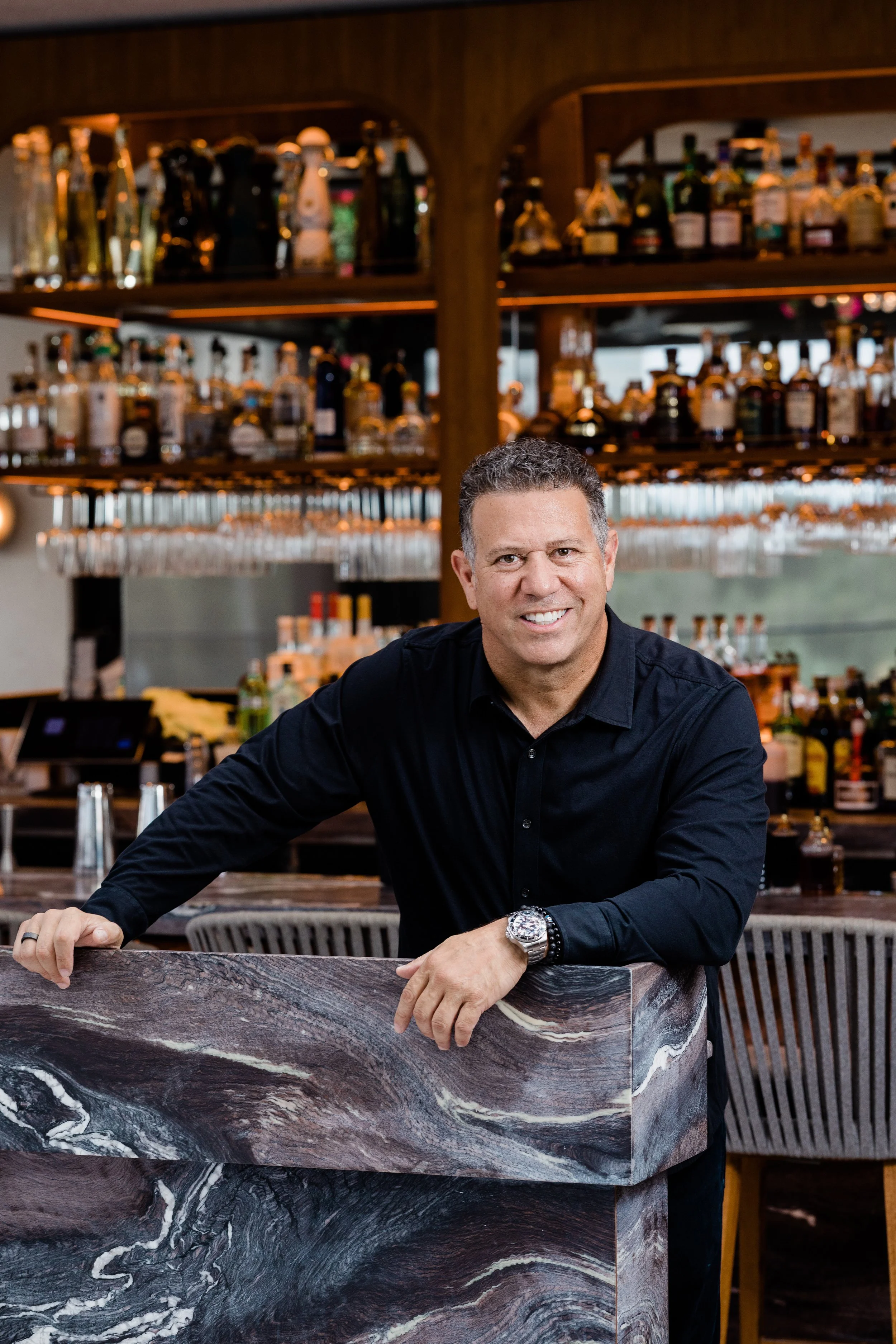 A man with curly dark hair smiling, dressed in a black shirt, sitting at a marble bar in a restaurant or bar setting with liquor bottles on shelves in the background.