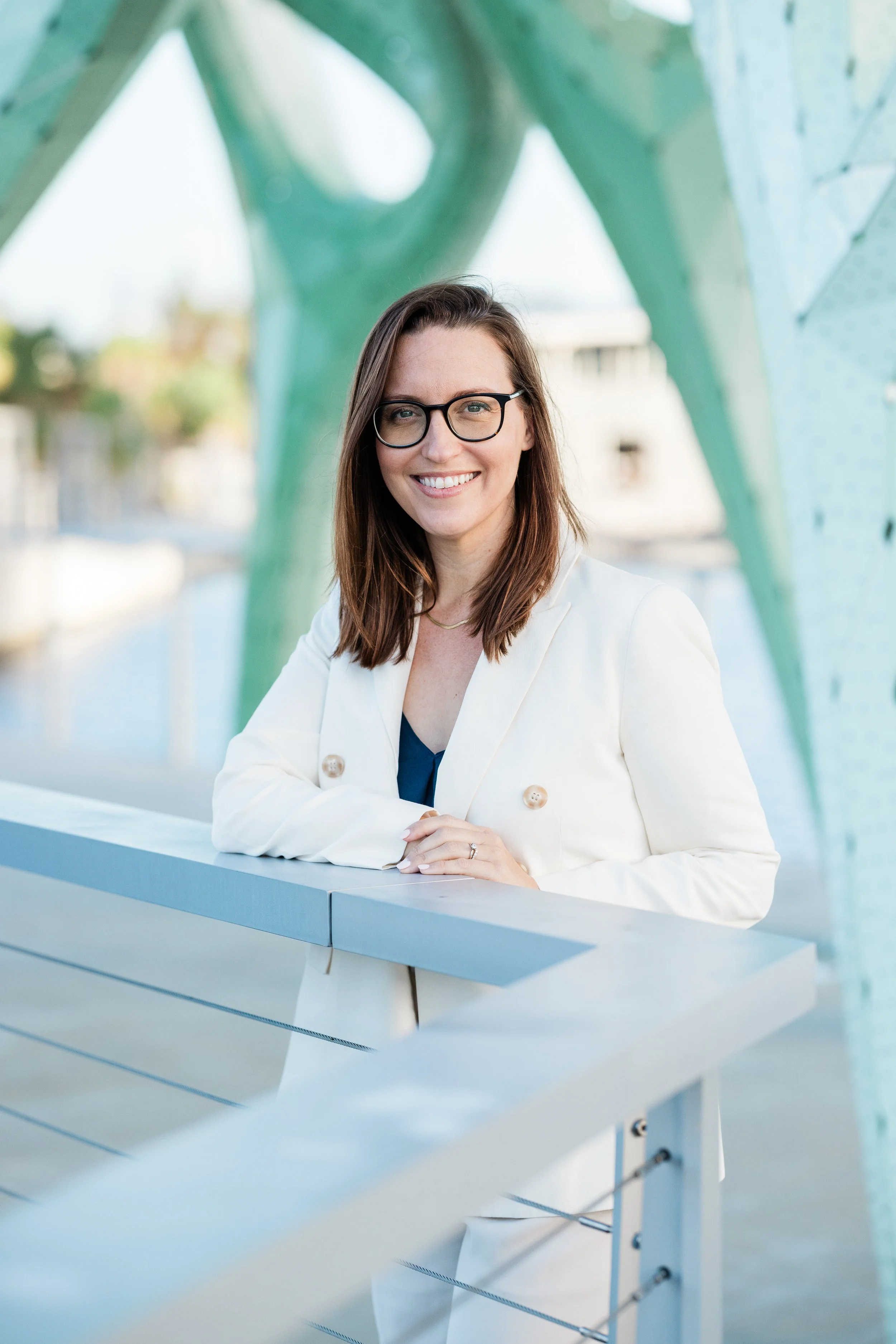 A woman with shoulder-length brown hair, glasses, and a white blazer smiling and leaning on a white railing outdoors, with modern green architectural structures and blurred cityscape in the background.
