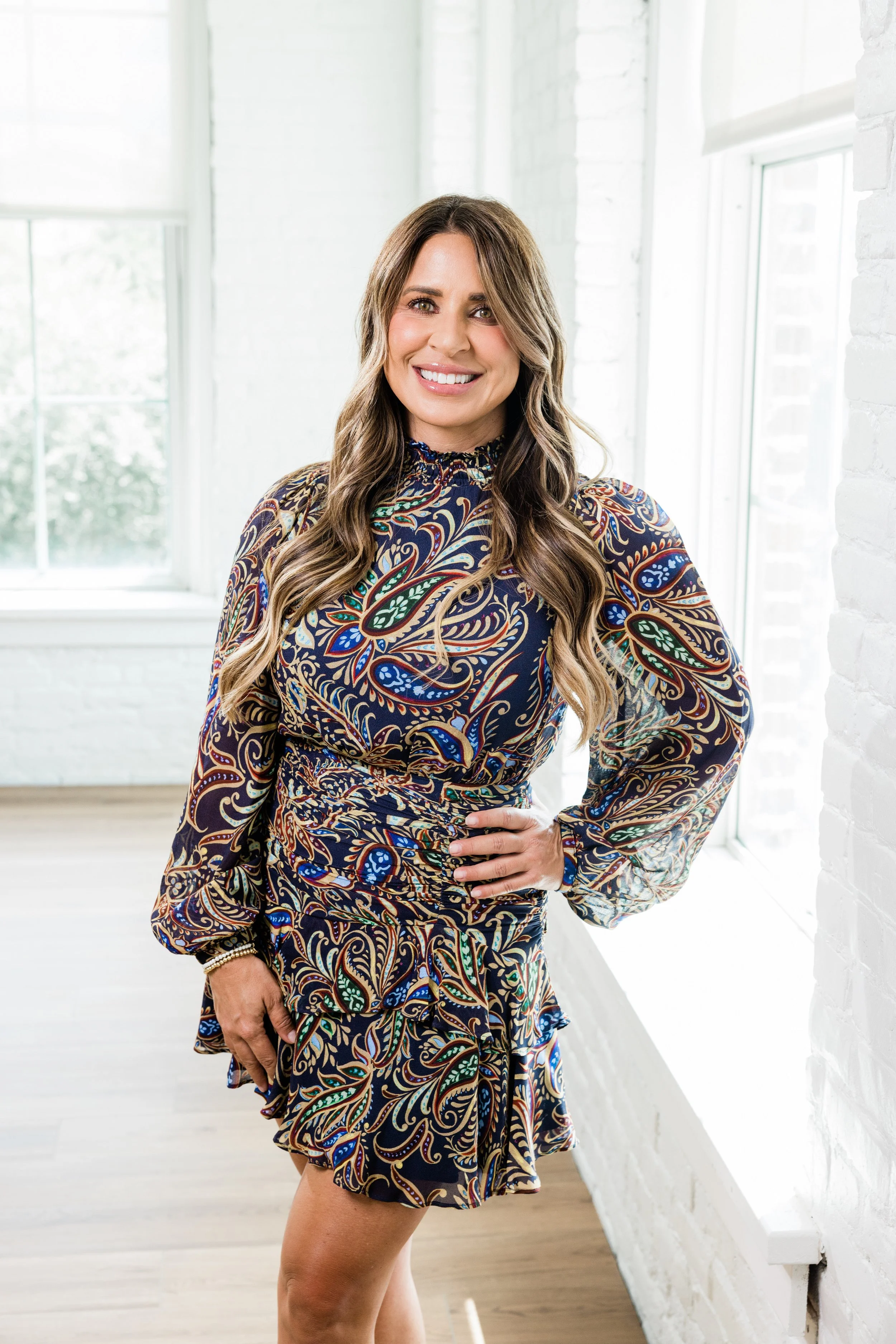 A woman with long, wavy hair smiling, wearing a colorful paisley patterned dress, standing in a bright white room with large windows and white brick walls.