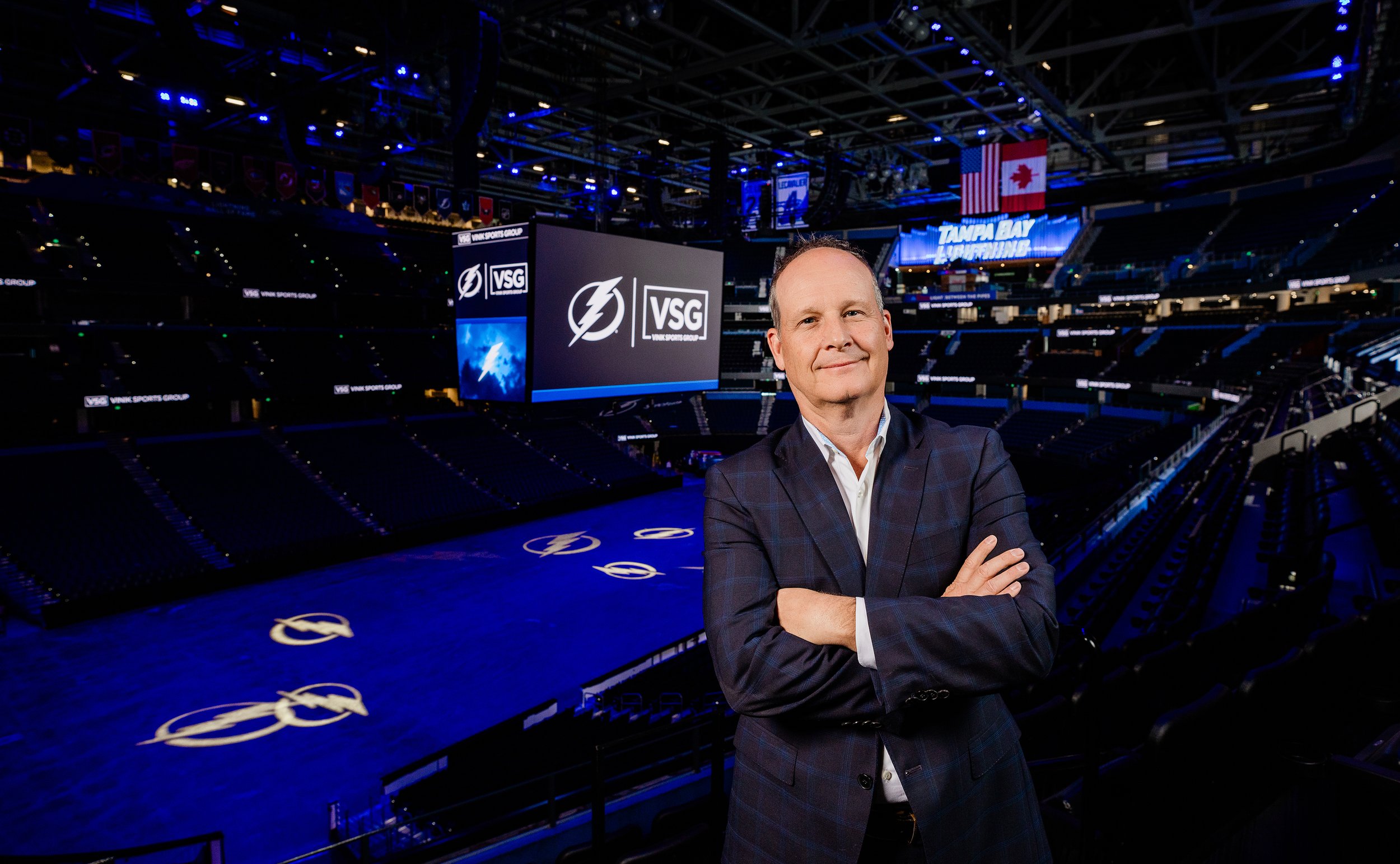 A man in a dark checkered suit with crossed arms standing inside an empty sports arena with large screens displaying 'VSG' logos and the Tampa Bay Lightning logo, with flags hanging from the ceiling.