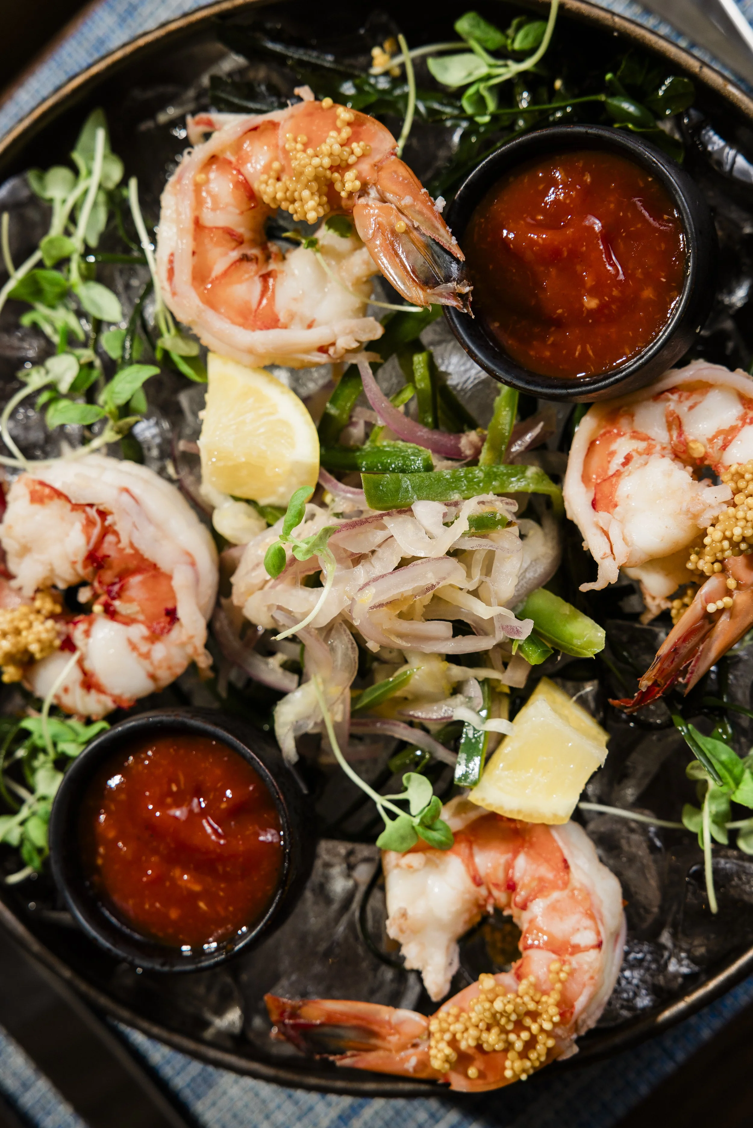 Close-up of a platter with cooked shrimp garnished with mustard seeds, lemon wedges, green herbs, and two small bowls of red dipping sauce.