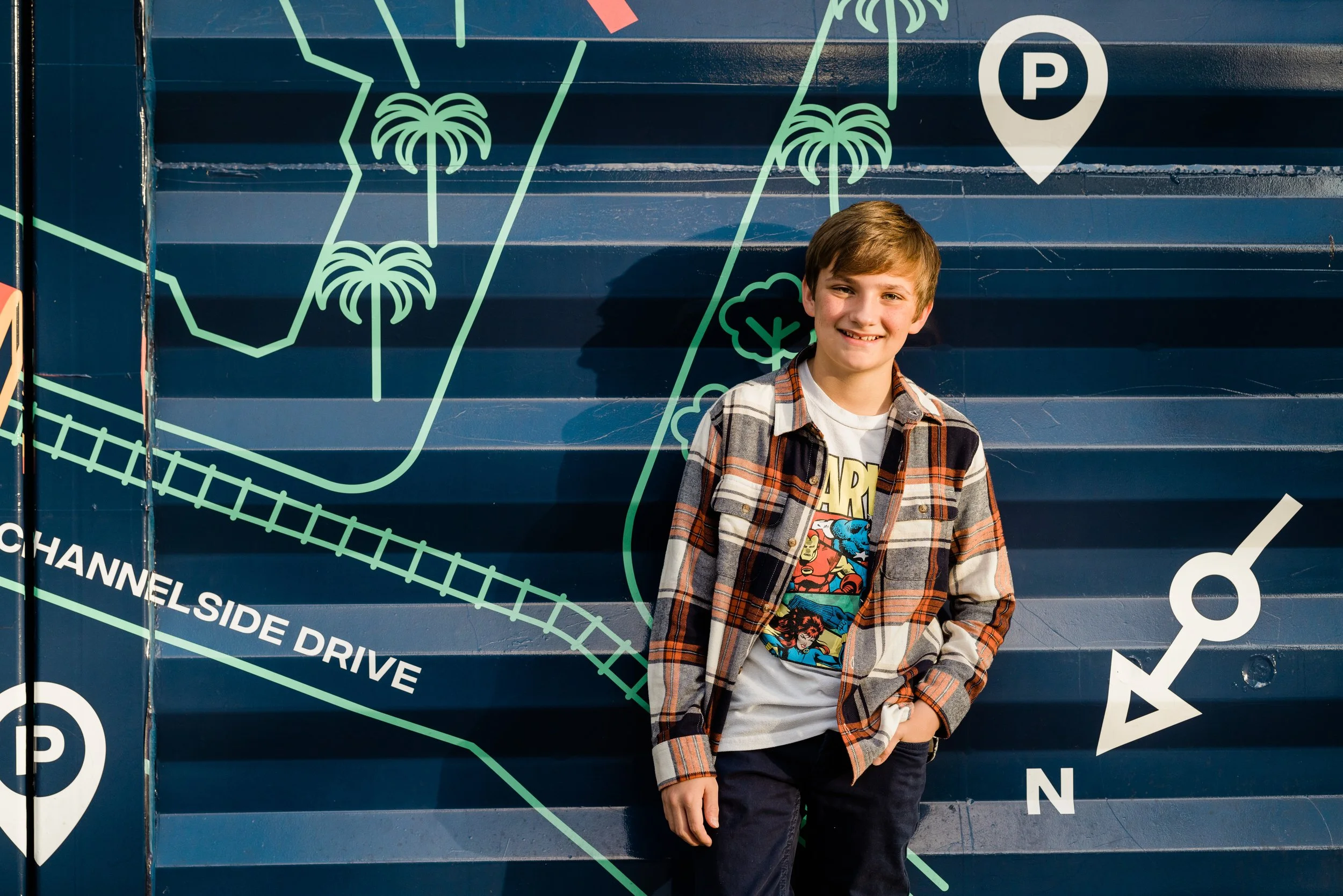A young boy with light brown hair, wearing a plaid shirt over a graphic T-shirt featuring Marvel superheroes, is standing against a dark blue wall with neon green line drawings of a palm tree, a cloud, and a parking icon, as well as white words inclu