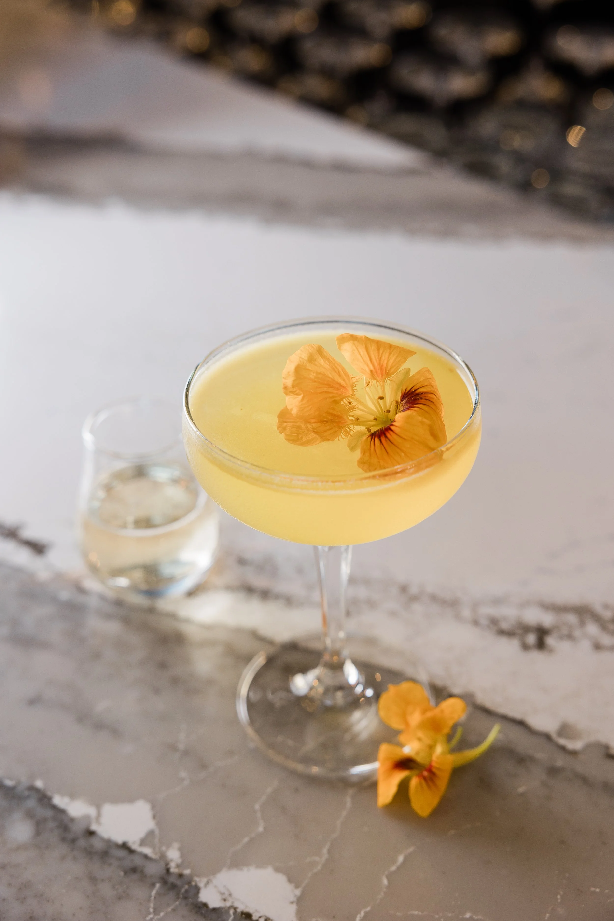 A cocktail glass filled with a pale yellow drink garnished with orange flower petals, placed on a marble surface with additional flower petals nearby.