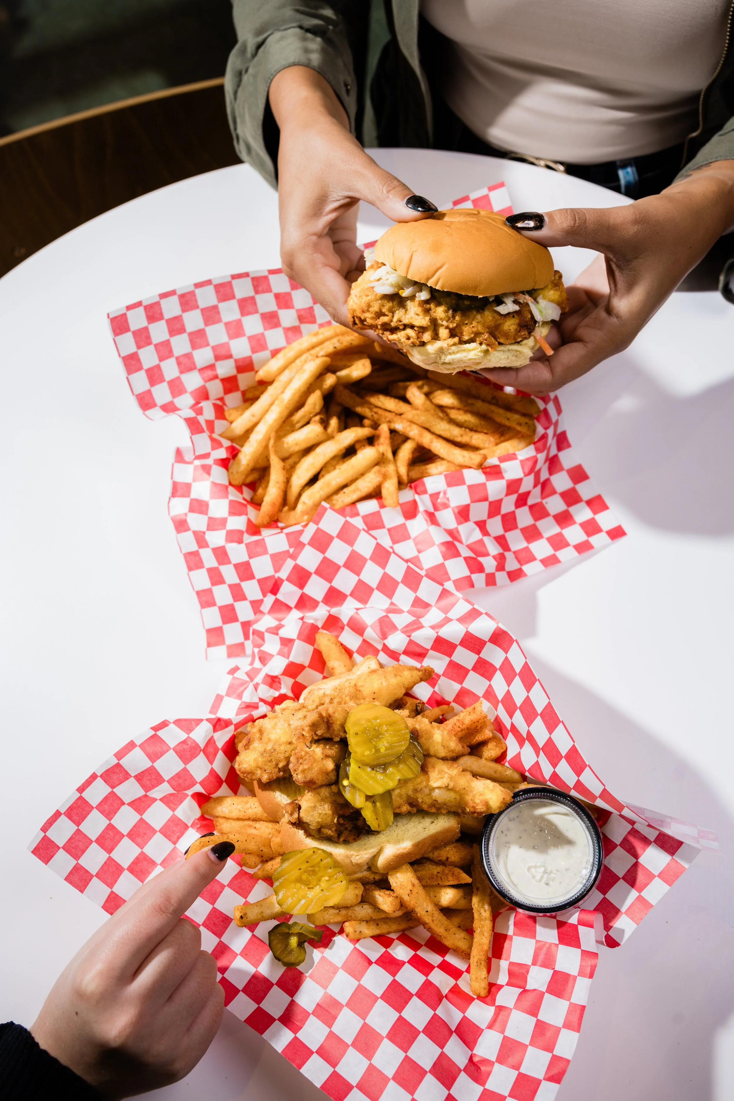 Hands holding a fried chicken sandwich with pickles, on a red and white checkered paper, with French fries on a white table, and a small cup of dipping sauce.