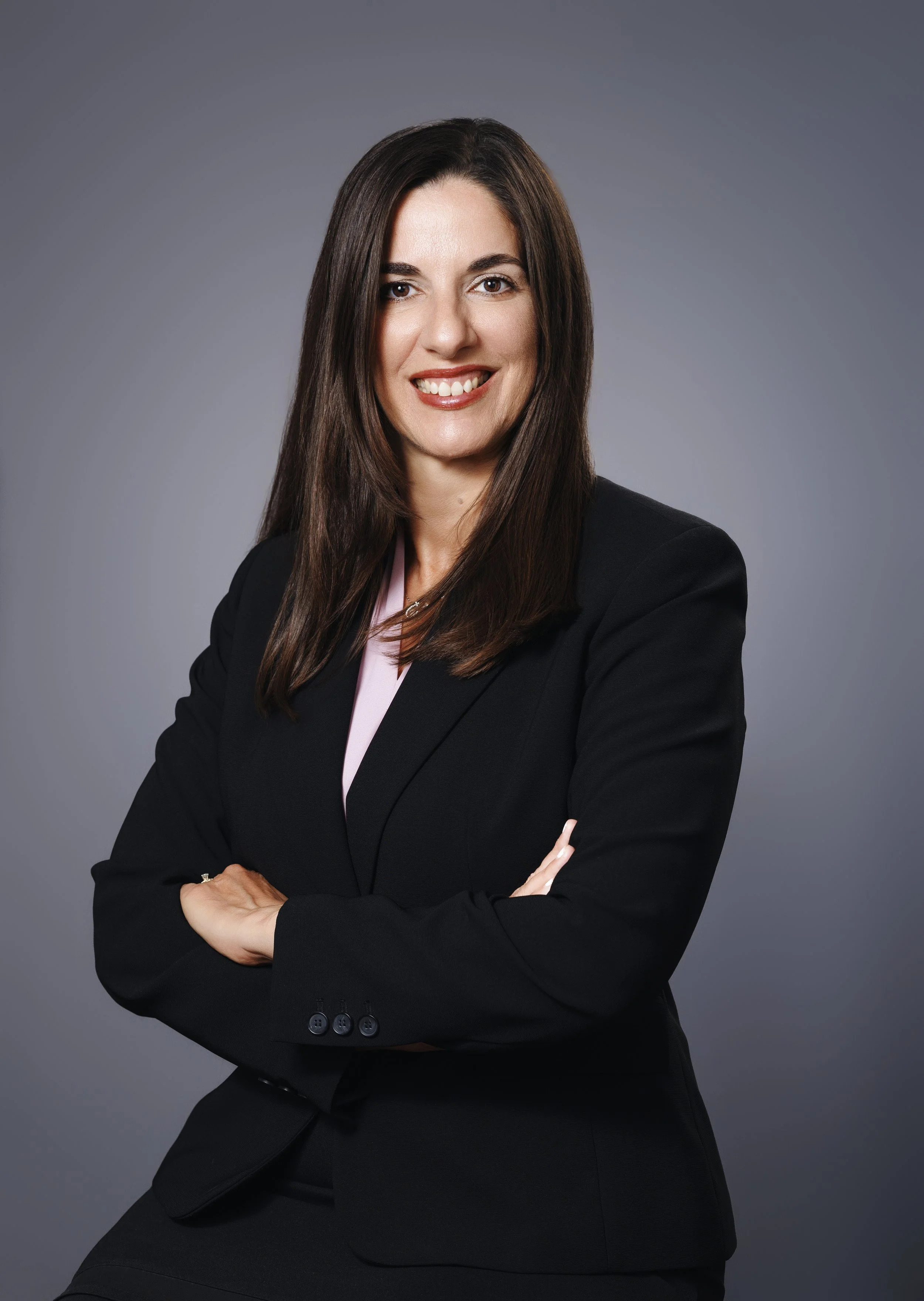 Professional woman with long brown hair, wearing a black blazer, smiling with arms crossed, against a plain gray background.