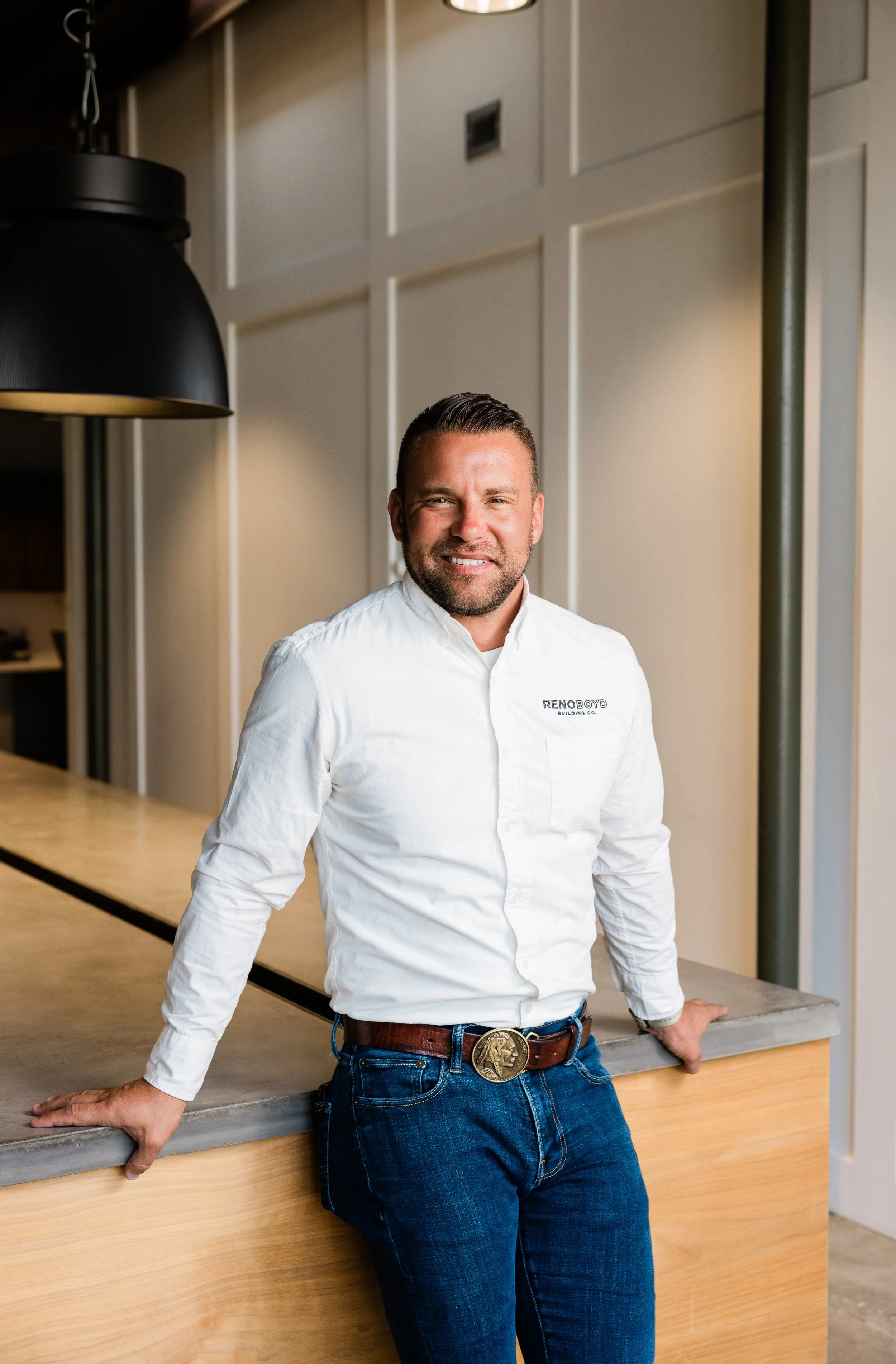Man wearing a white shirt with a logo that says 'Renoboyd Building Co.' leaning on a counter in a modern, well-lit room with beige paneled walls and black and gold pendant lighting.
