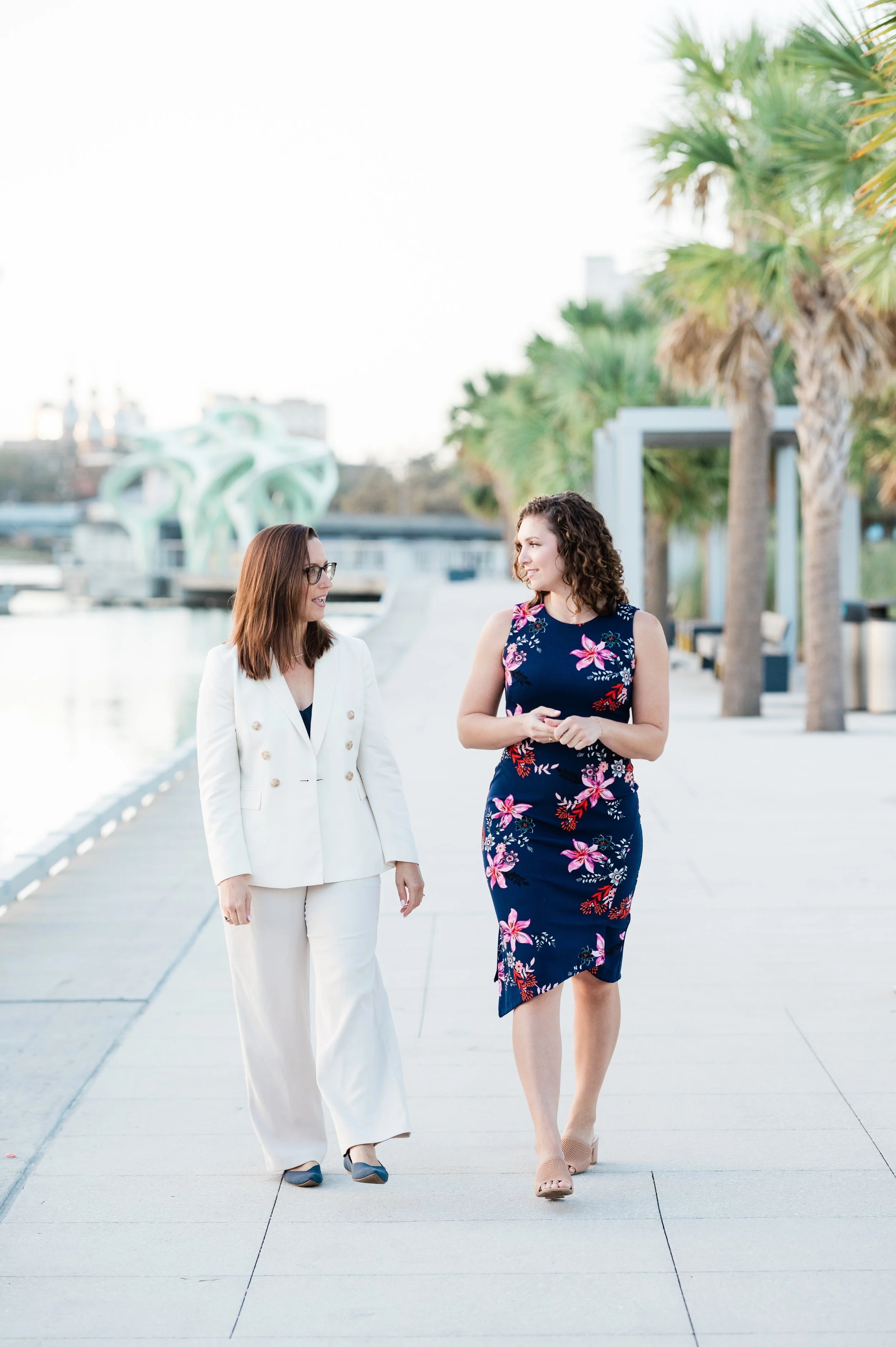 Two women are walking and talking outdoors near palm trees with a modern sculpture and cityscape in the background.