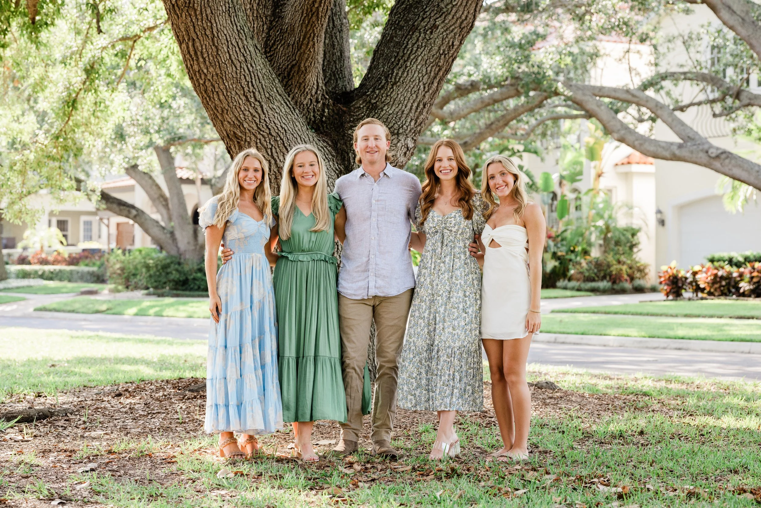 A group of five young adults standing arm in arm in front of a large tree in a suburban neighborhood, smiling at the camera.
