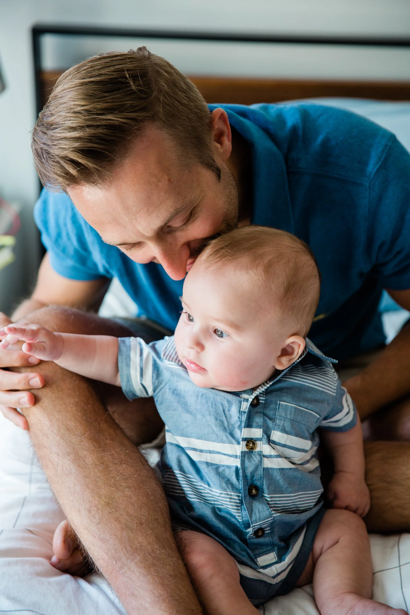 A man is sitting on a bed, holding a young child, and kissing the child's forehead. The child is sitting and looking to the side, wearing a blue and white striped shirt.