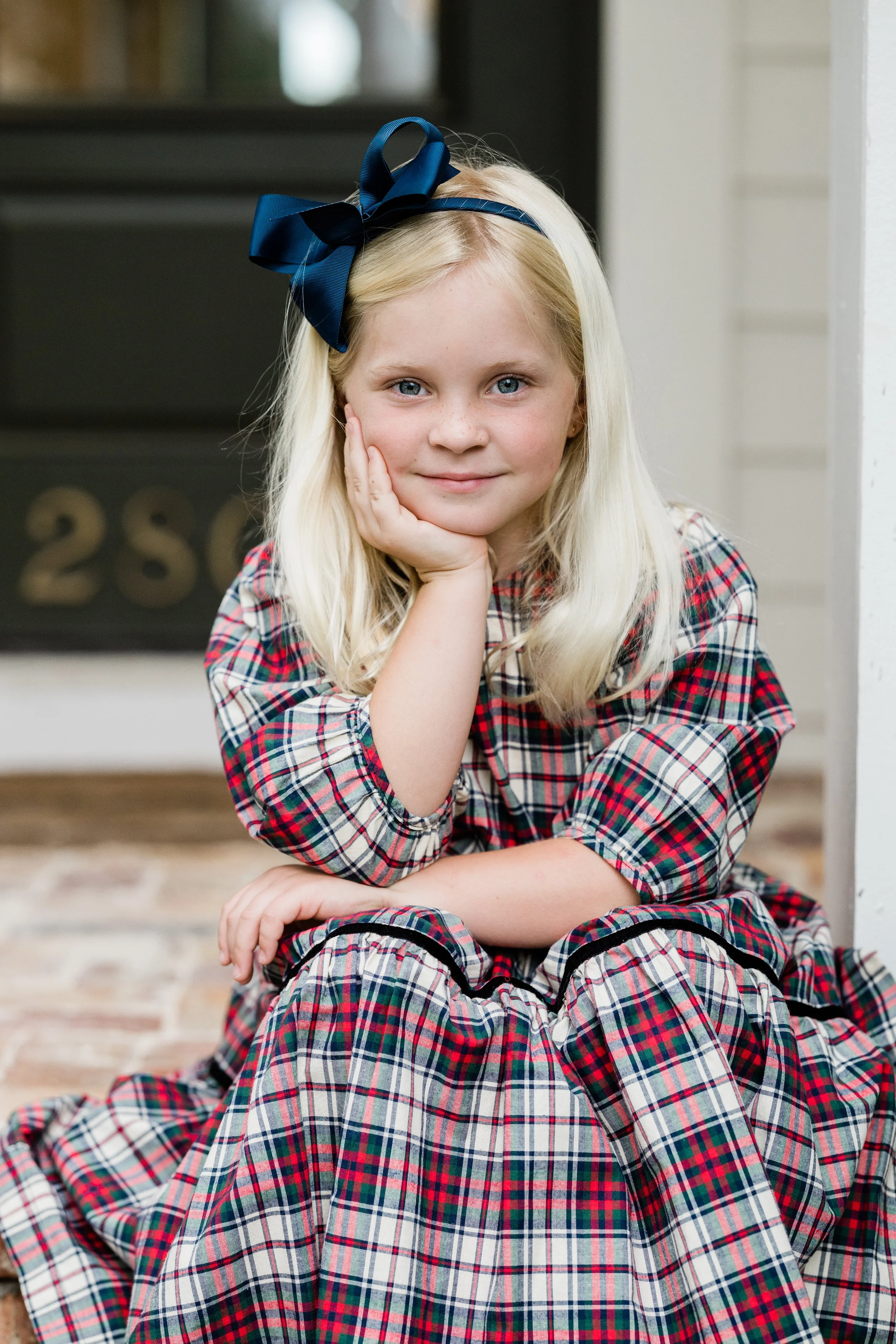 A young girl with blonde hair and blue eyes sitting on a front stoop, resting her head on her hand, wearing a plaid dress and a large navy blue bow in her hair.