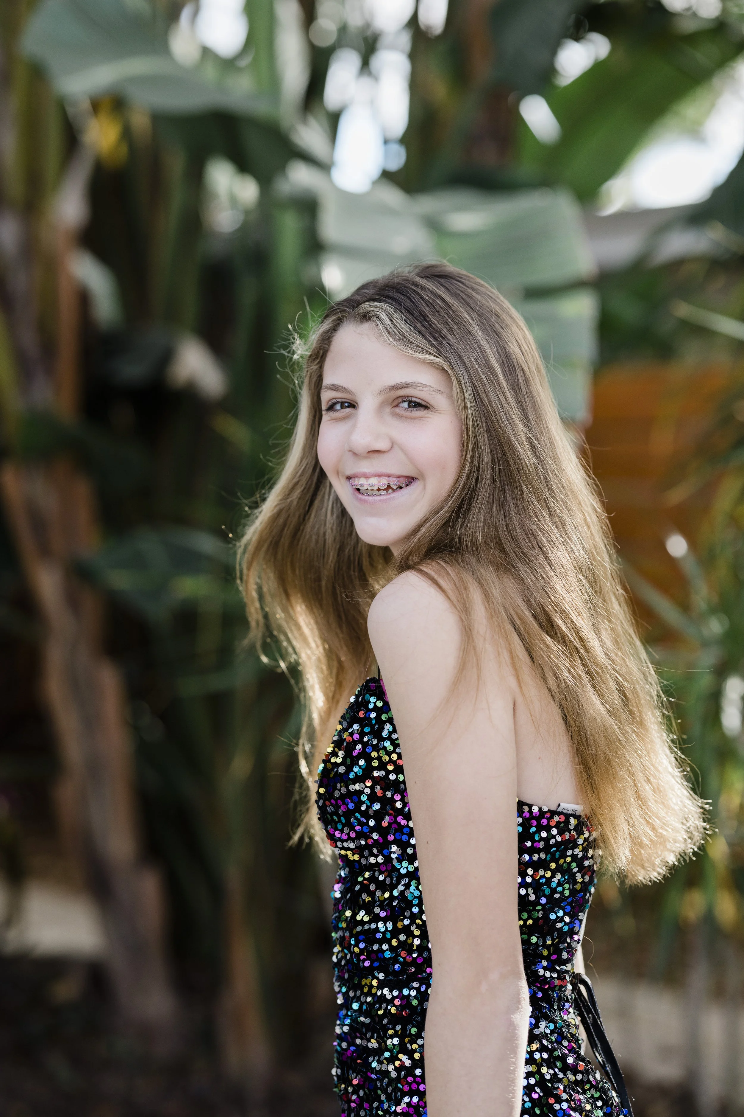 A young girl with long, light brown hair smiling outdoors. She is wearing a black dress sequined with multicolored shiny dots. The background features large green leaves.