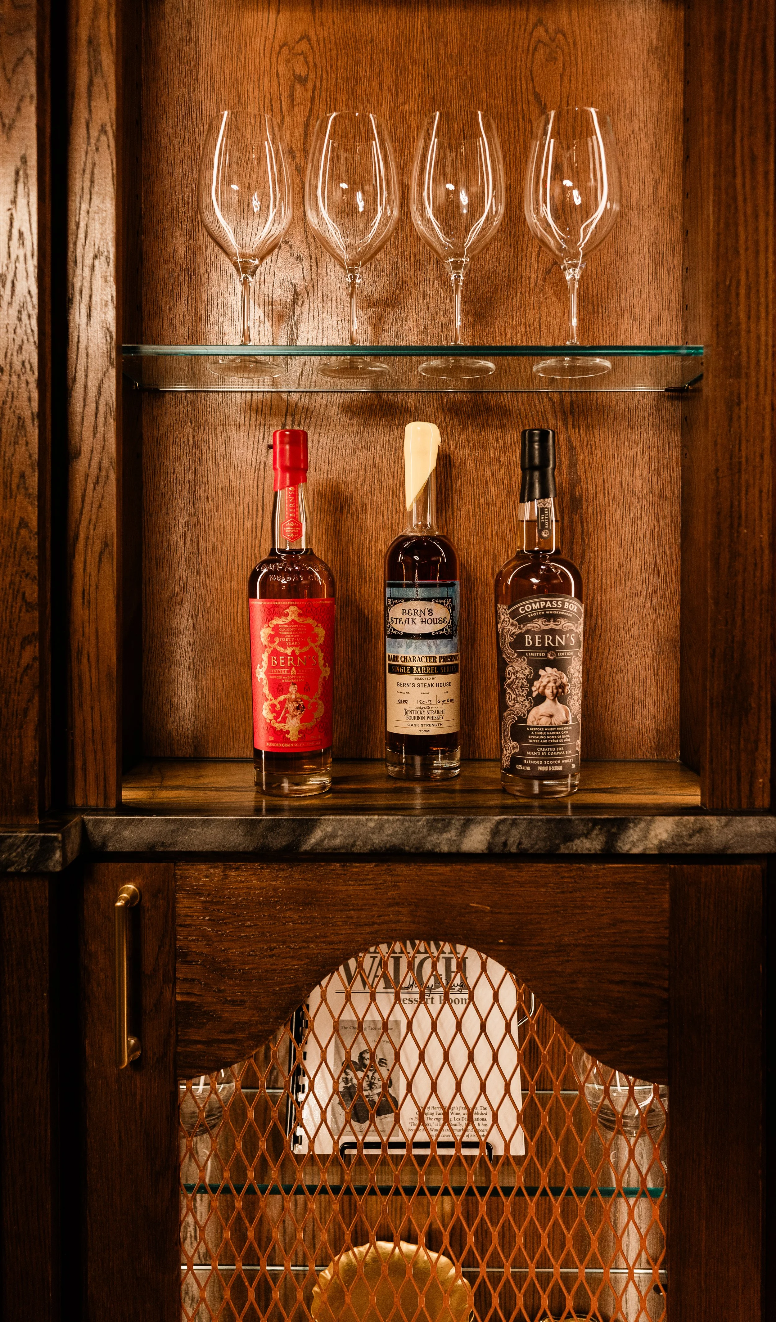 Wooden cabinet with three empty wine glasses on the top shelf and three bottles of whiskey on the middle shelf, behind a metal mesh door.