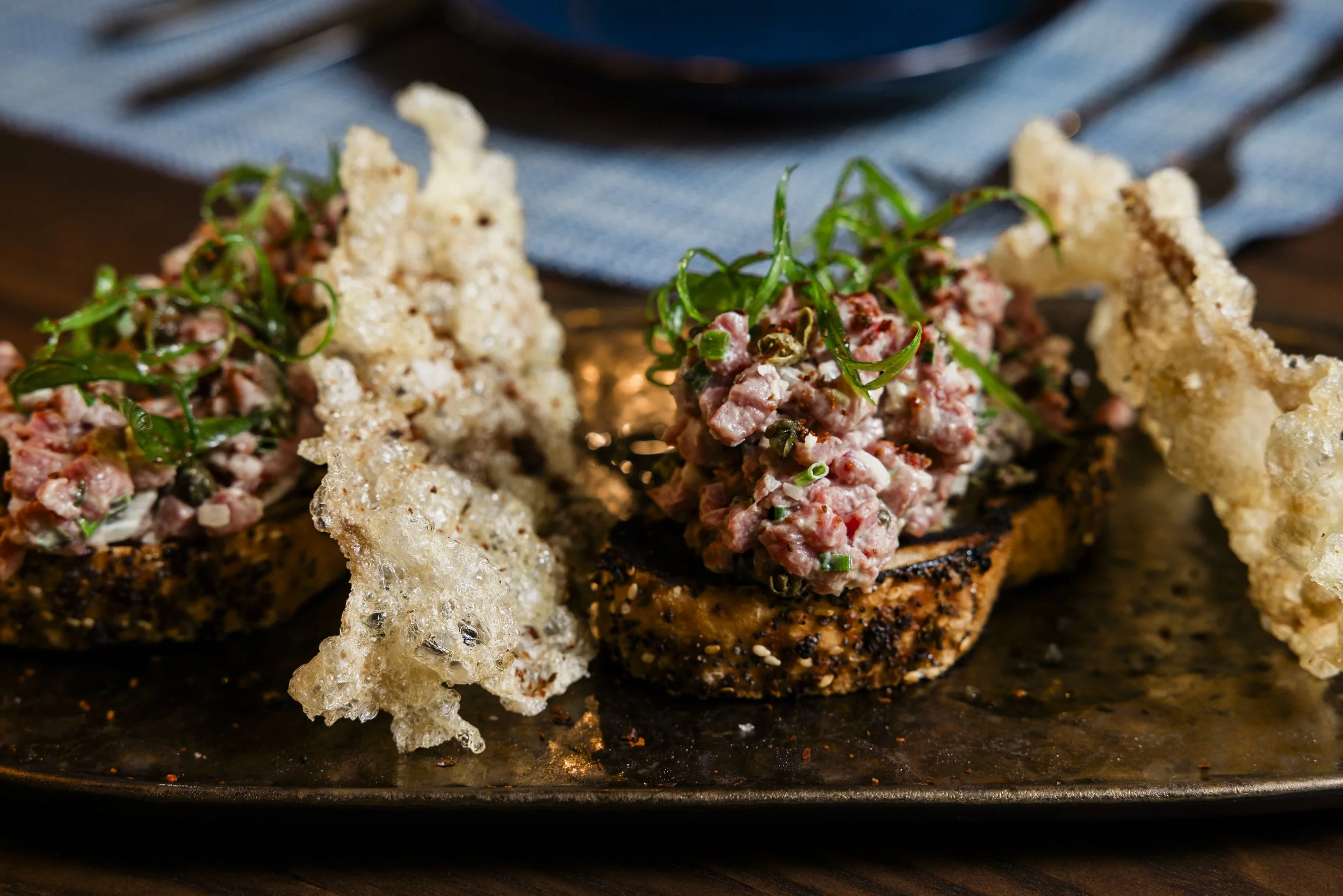 Close-up of gourmet canapé with minced meat topping and green garnish, served on crispy bread with parmesan cheese and crispy rice crackers on a dark serving tray.