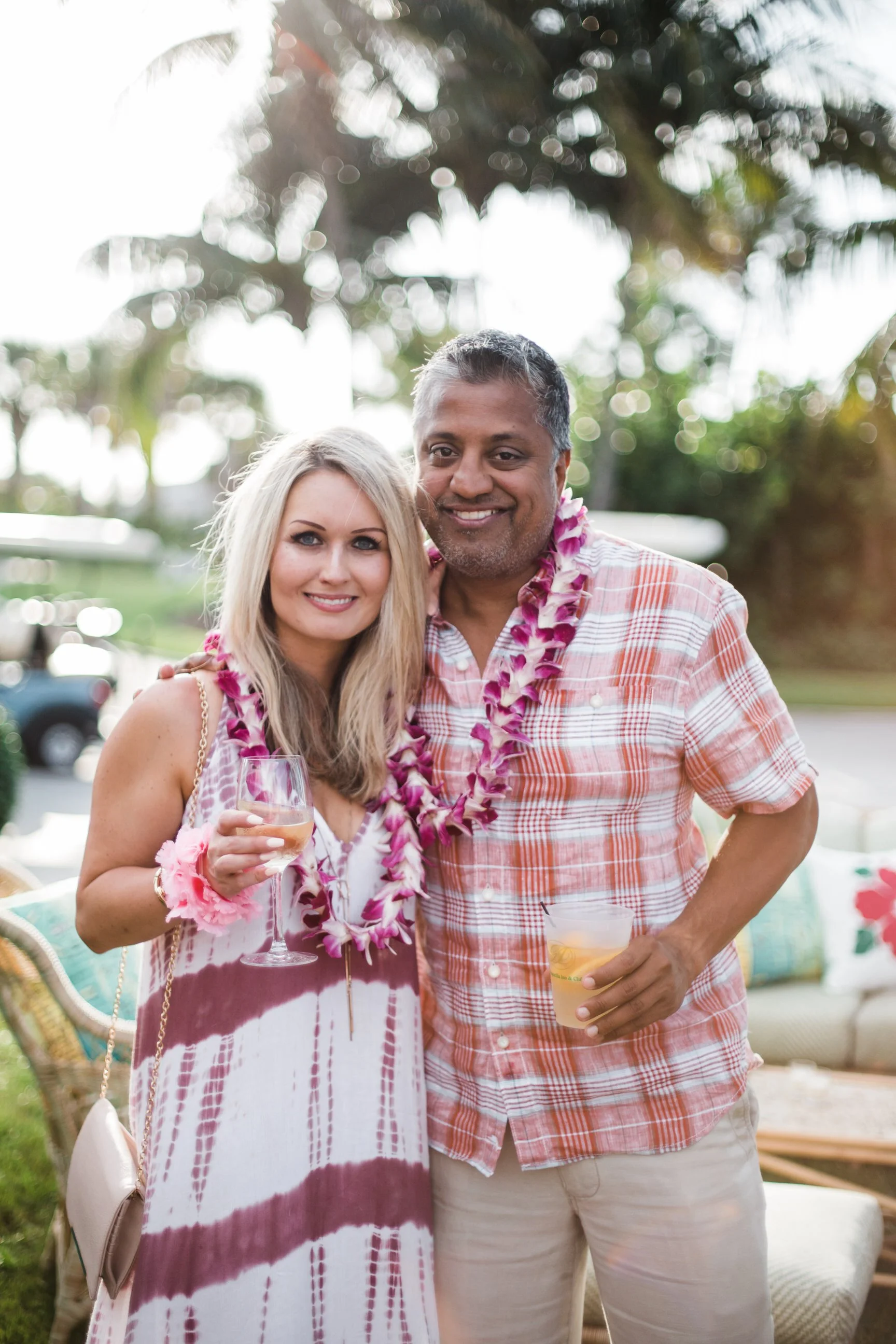 A man and a woman are standing together outdoors, both wearing leis around their necks. The woman is holding a glass of wine, and the man is holding a drink in a plastic cup. They are smiling at the camera, and the background features blurred trees a