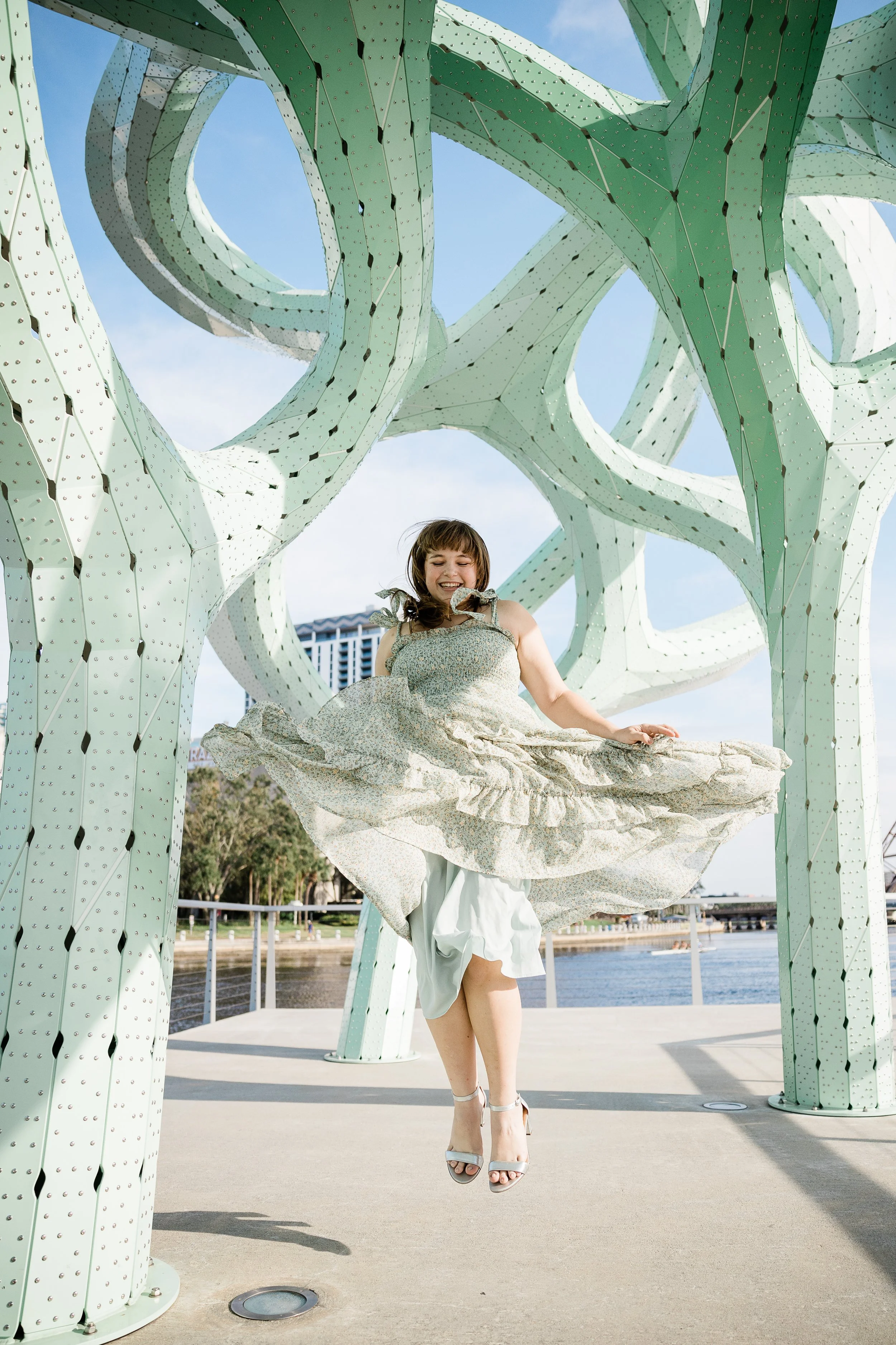 A woman in a light-colored dress is sitting on a suspended platform under a large, artistic metal sculpture structure with green, white, and gray colors. She appears to be smiling and happy, with her eyes closed, and is wearing high-heeled shoes. The