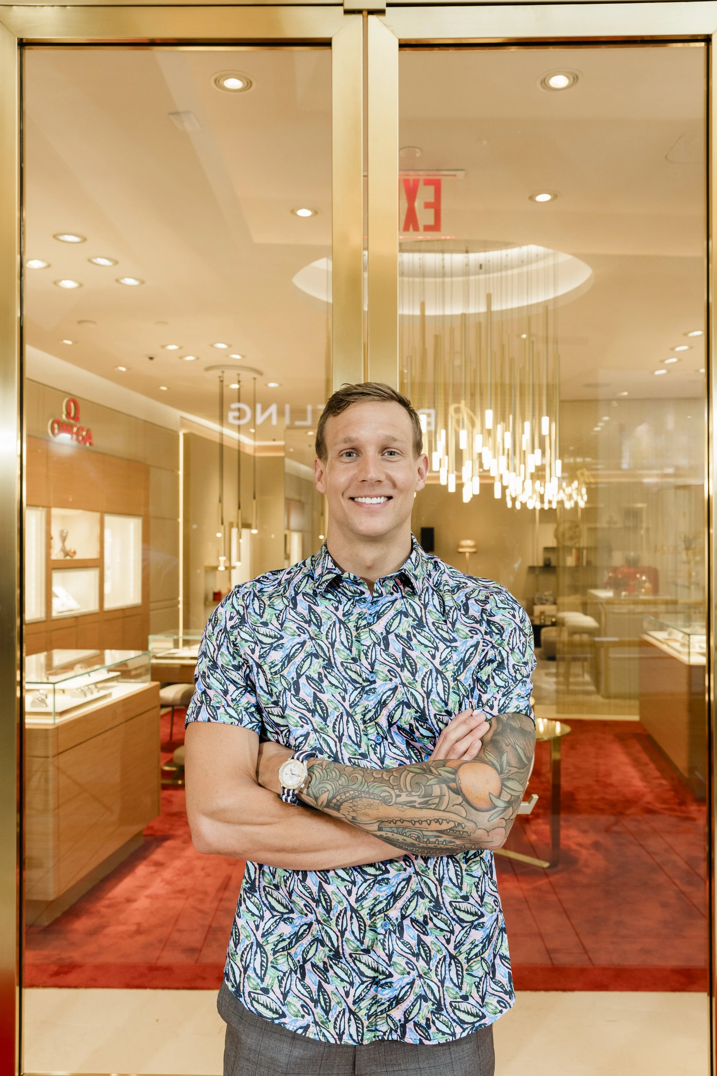 A man with short brown hair and tattooed arms, smiling with arms crossed, standing in front of a glass door in a shopping mall or jewelry store.