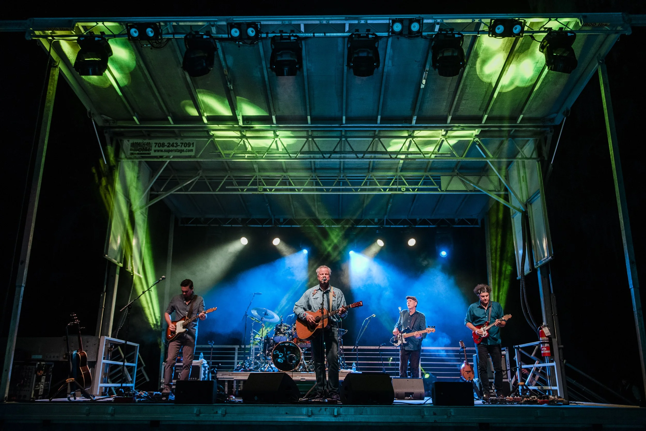 A live band performing on an outdoor stage at night, with musicians playing guitars, drums, and singing, under colorful stage lighting.