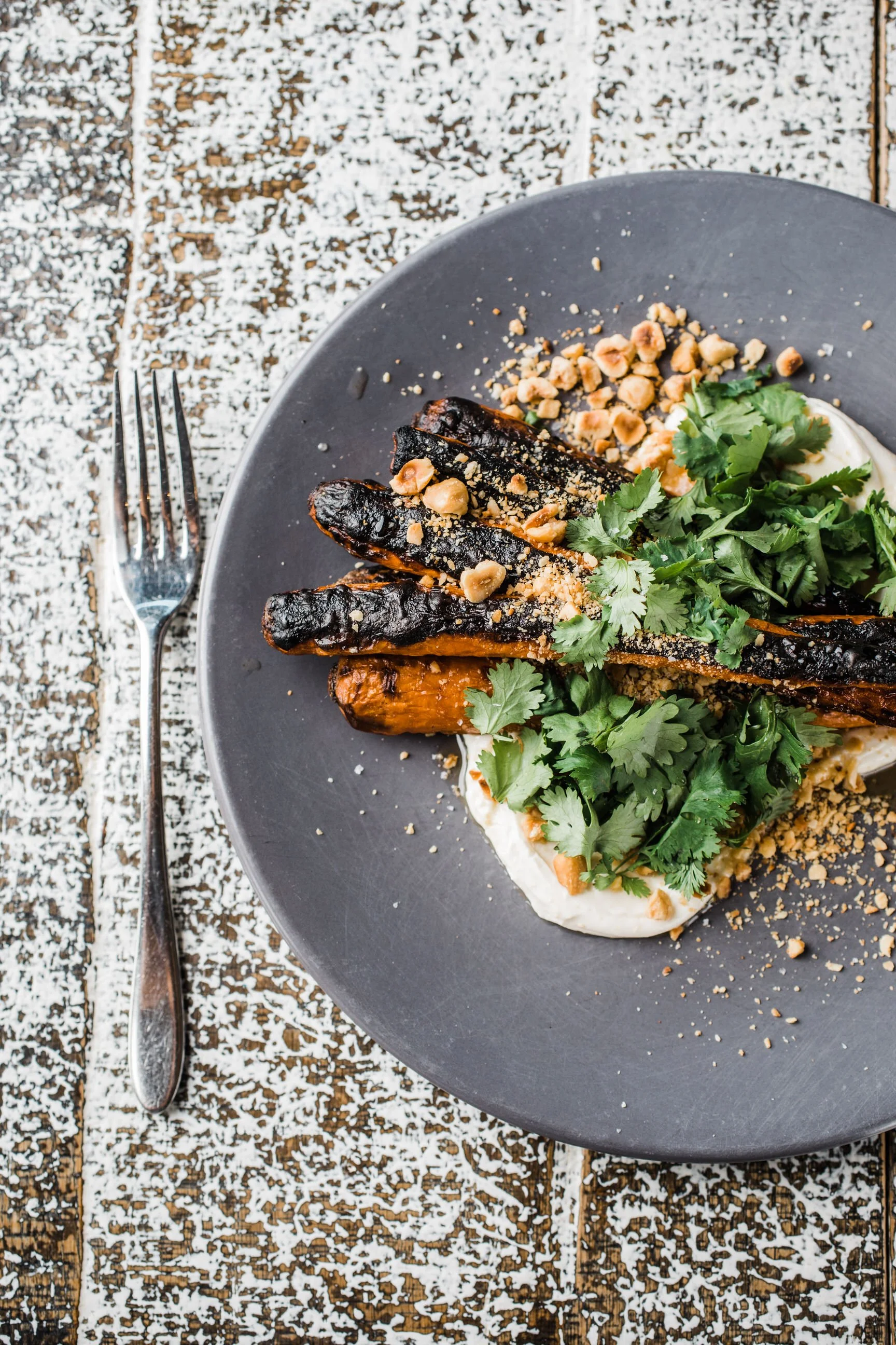 A plate of roasted carrots topped with chopped nuts and fresh cilantro, served on a bed of creamy white sauce with crushed nuts sprinkled around, on a textured wooden surface with powdered sugar or flour.