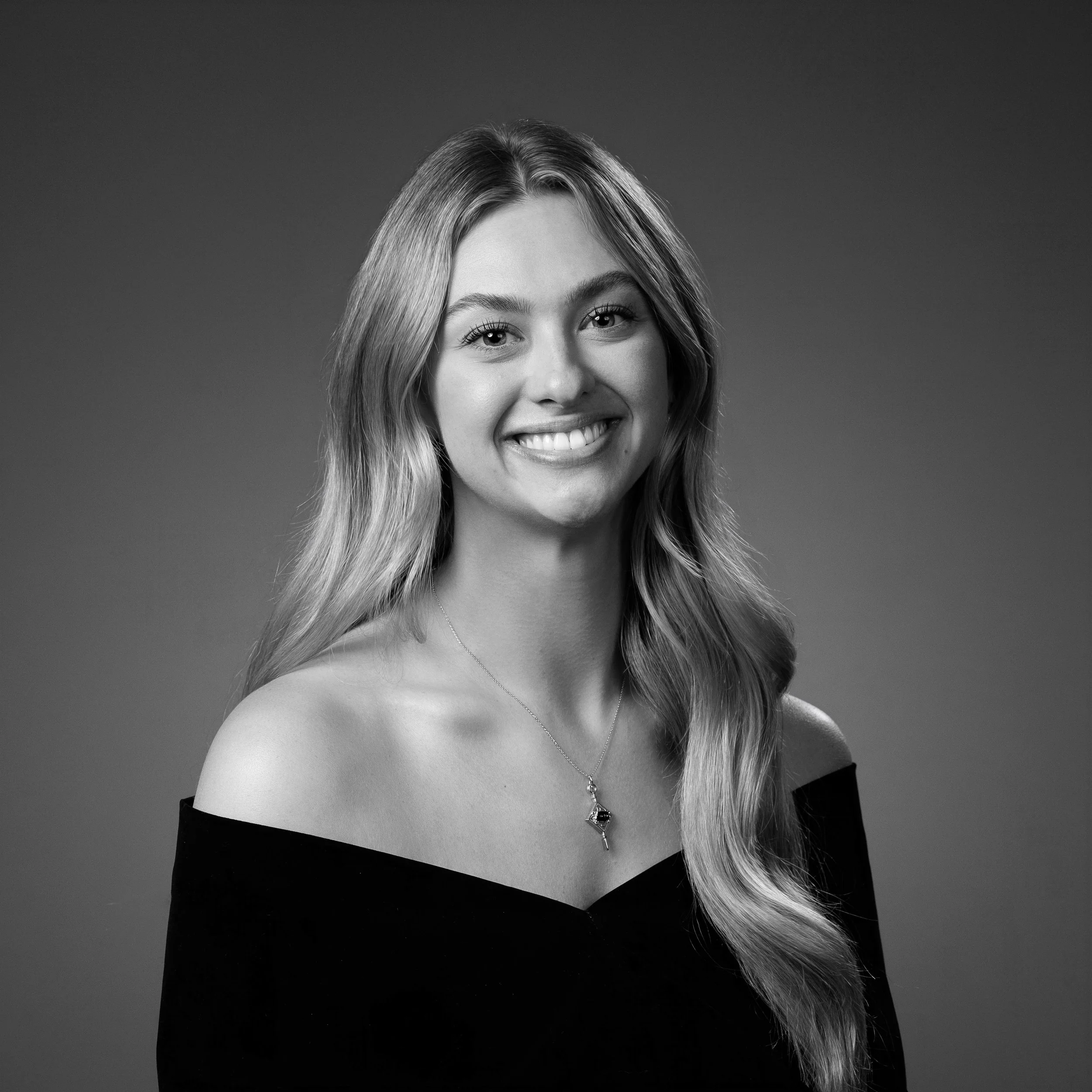 Black and white portrait of a woman with long hair, smiling, wearing an off-the-shoulder top, necklace, and earrings.