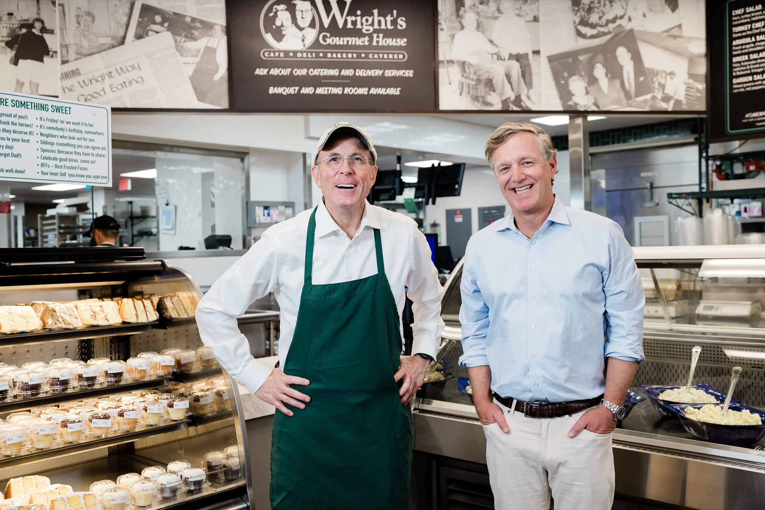 Two men smiling inside Wright's Gourmet House, a deli and bakery, standing in front of display cases filled with desserts and food items.
