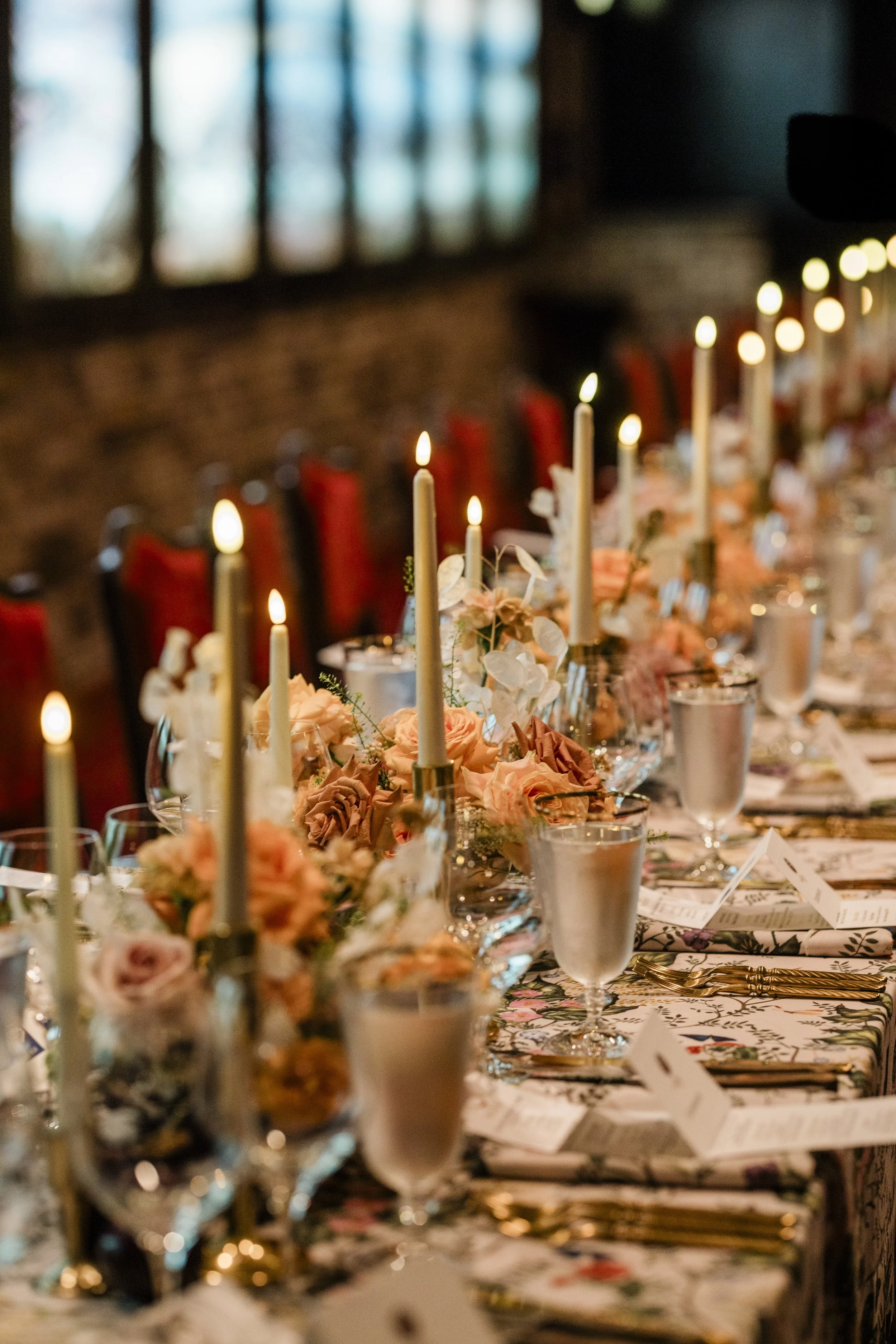 A long dining table decorated with pink and peach flowers, lit candles, and floral-patterned tablecloths, set for a formal Winefest event in a room with brick walls and stained glass windows. Bern's Rhone River photographs in background.